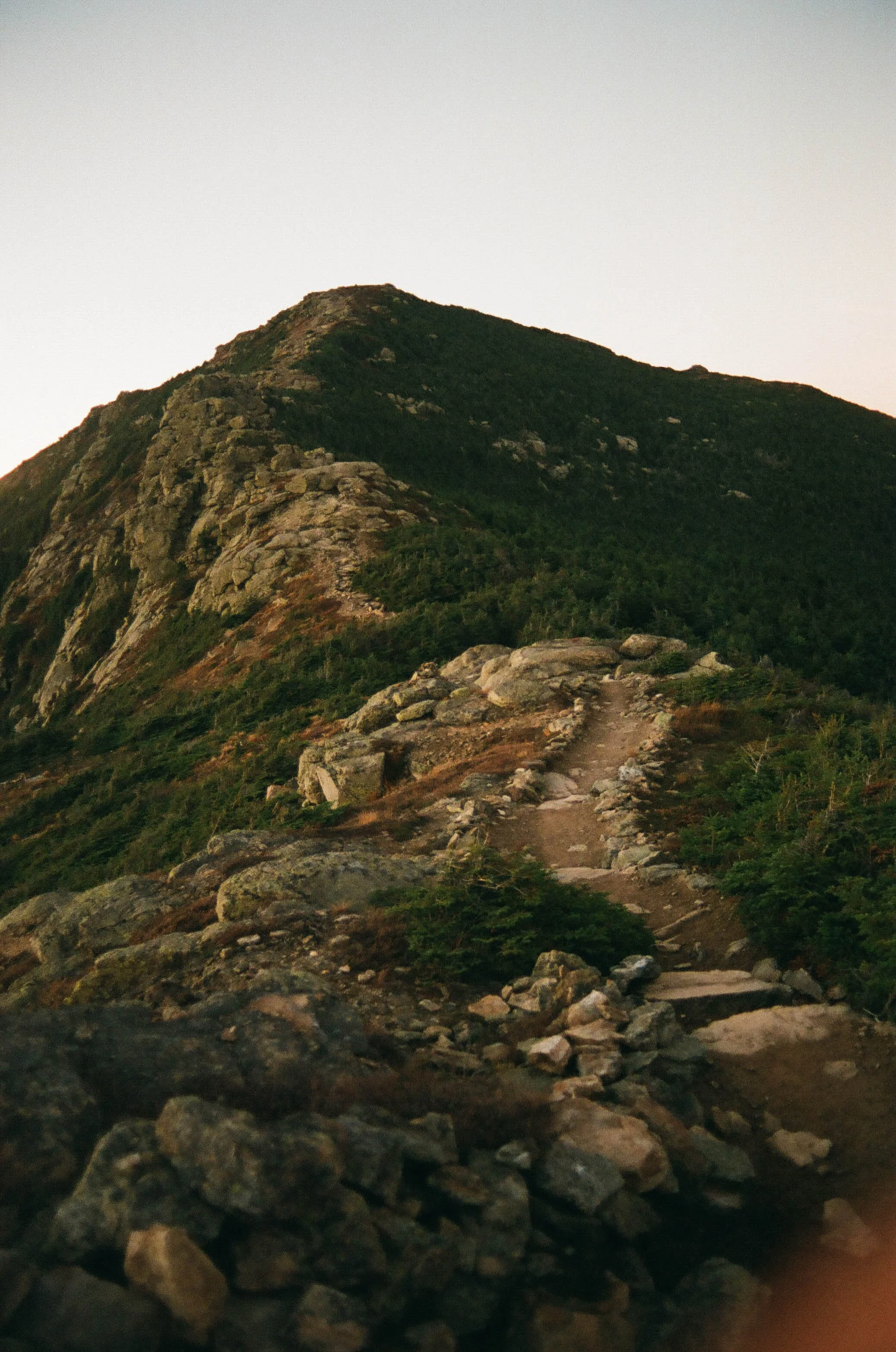 A mountain with a rocky trail leading up its slope, surrounded by green shrubbery and trees.