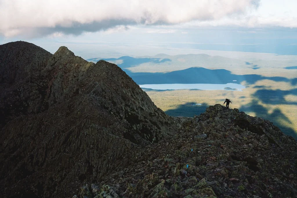 Hiker climbing rocky mountain ridge with a scenic view of mountains, lakes, and clouds in the distance.
