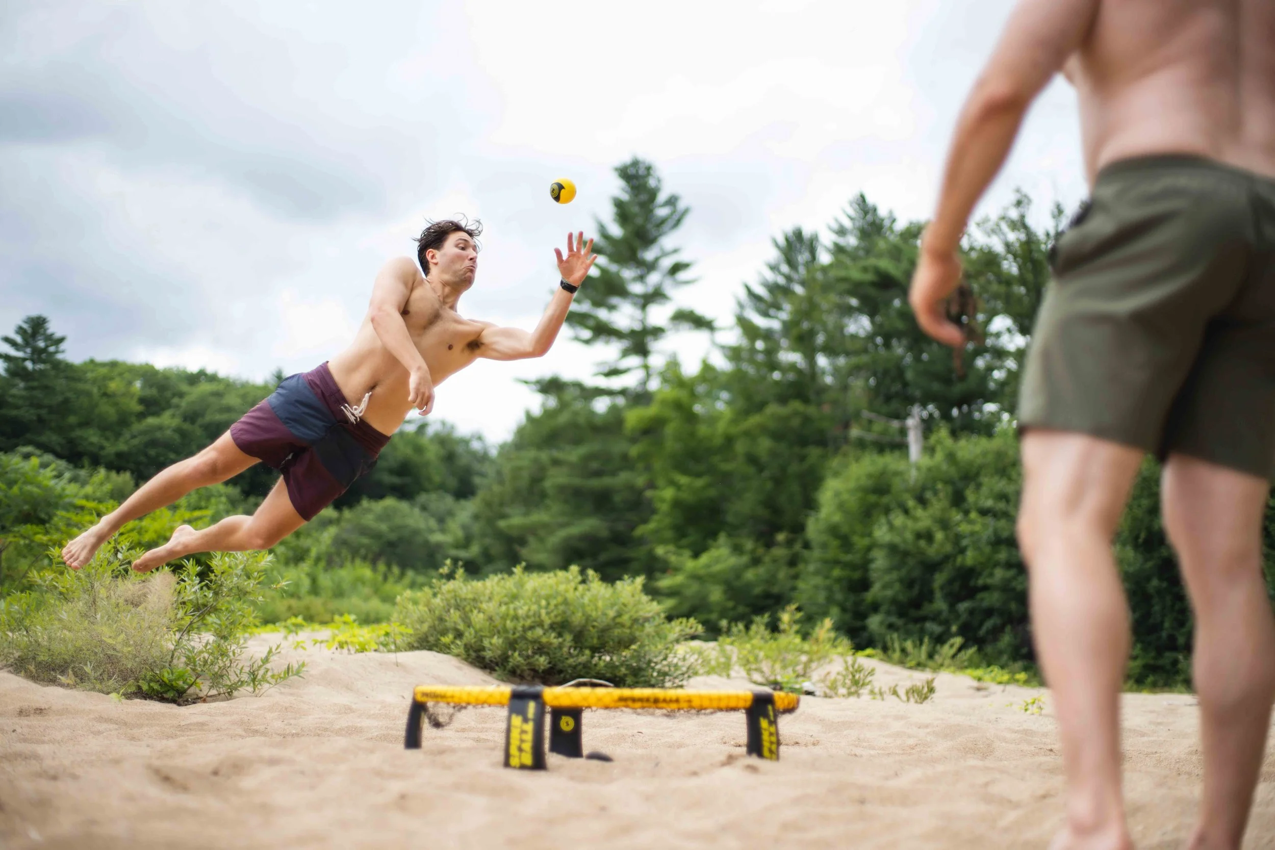 A young man dives mid-air to make a Spikeball play on a scenic river beach.