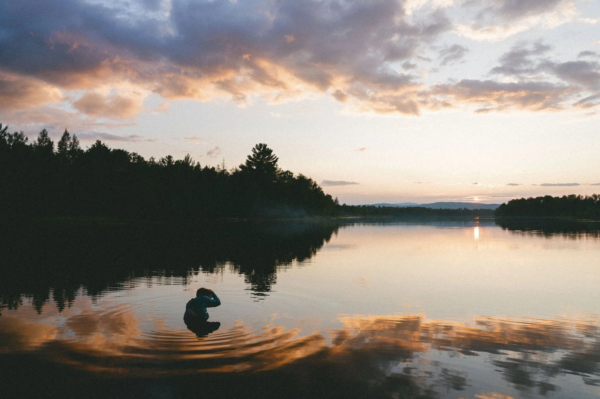 A person kneeling in a calm lake during sunset, surrounded by trees and reflecting colorful clouds on the water.
