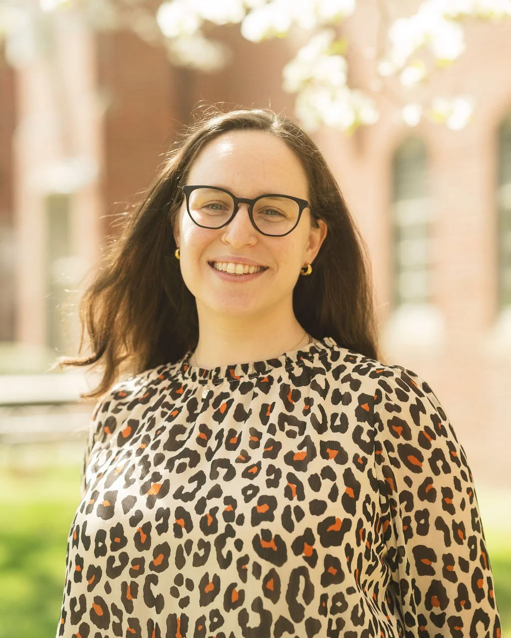 A woman with long brown hair, glasses, and gold hoop earrings smiling outdoors in sunlight, wearing a leopard print blouse.