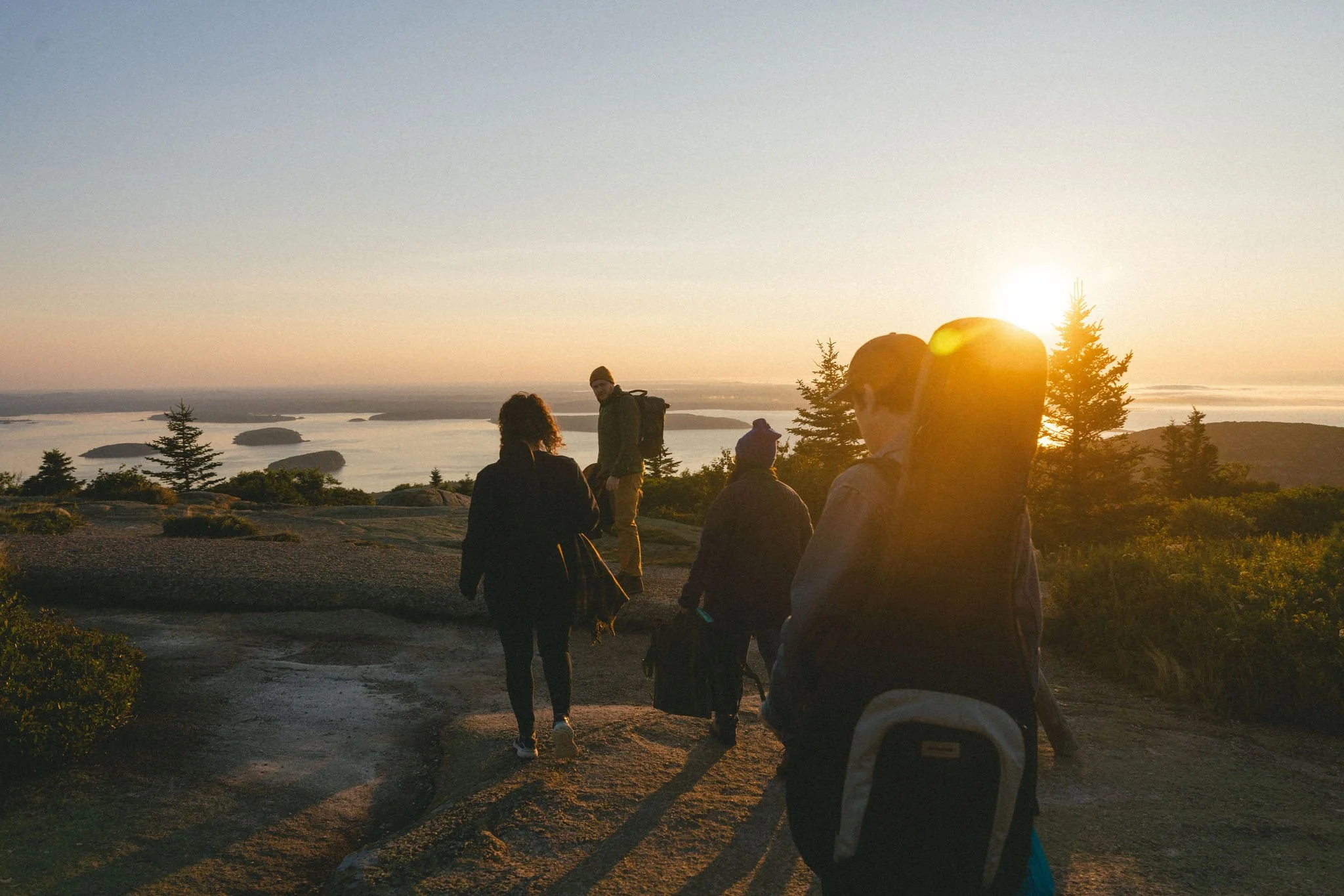 Group of people hiking at sunset on a rocky trail with trees and water in the background.