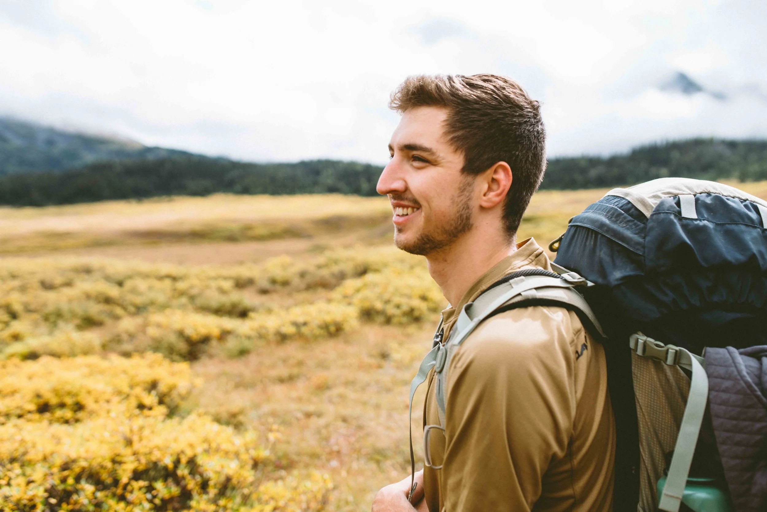 Young man hiking outdoors with a backpack, smiling, in a natural landscape with hills and yellow foliage.