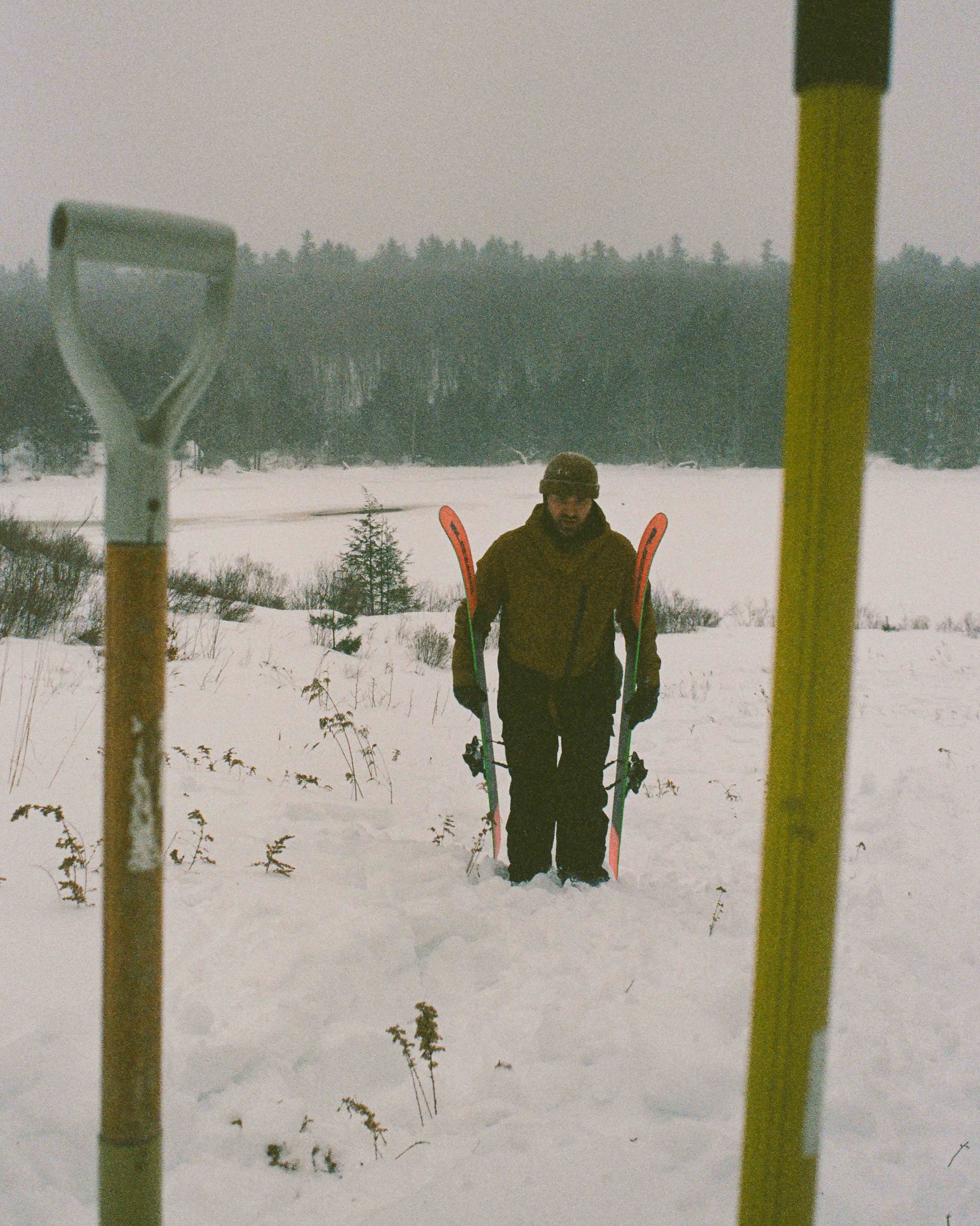A person standing in snow with skis, wearing a helmet and brown jacket, viewed from behind between two yellow skidoo flags, with a snowy landscape and trees in the background.