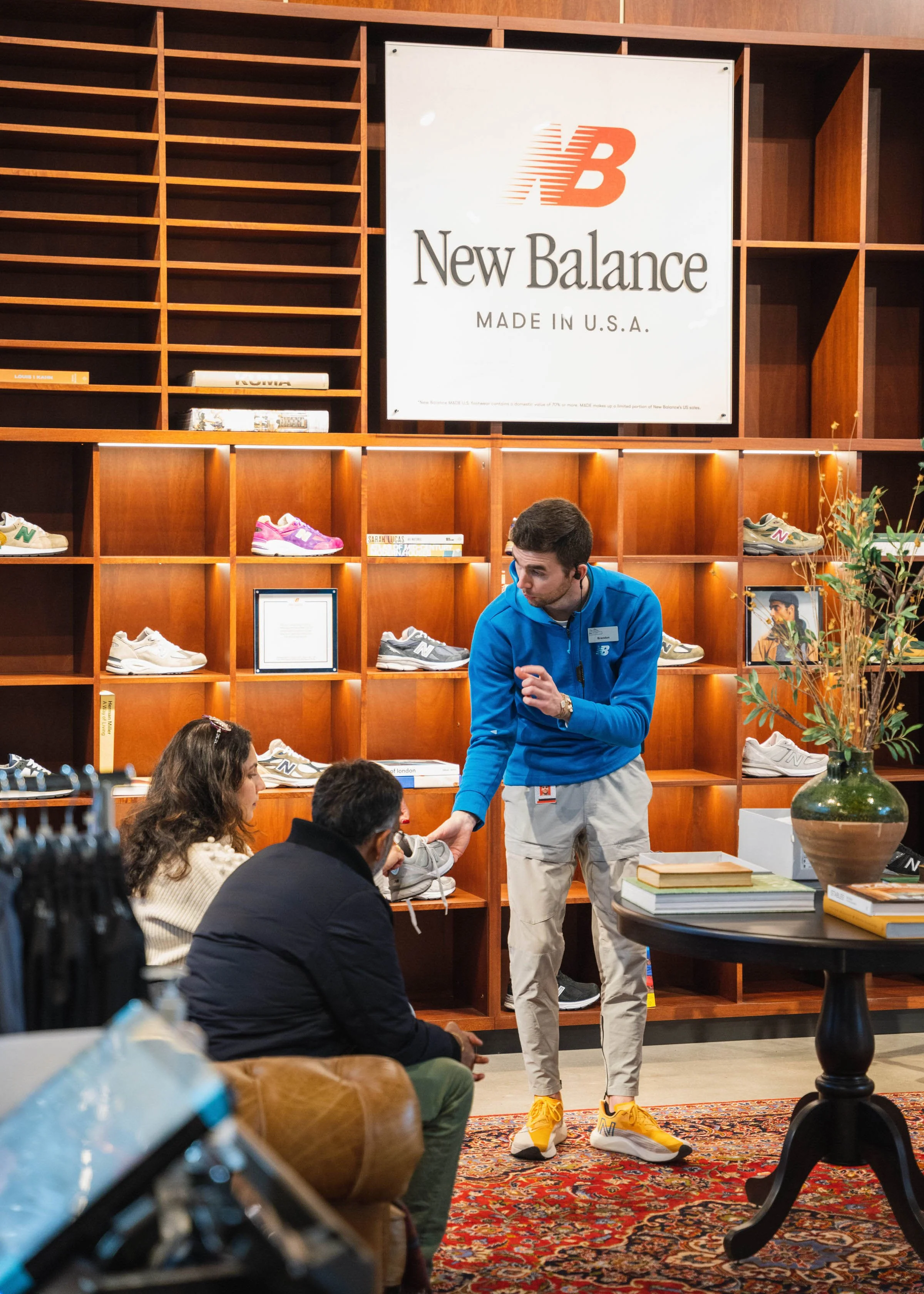 A New Balance employee showing a sneaker to two customers inside a store with wooden shelves displaying various sneakers and a large New Balance sign on the wall.