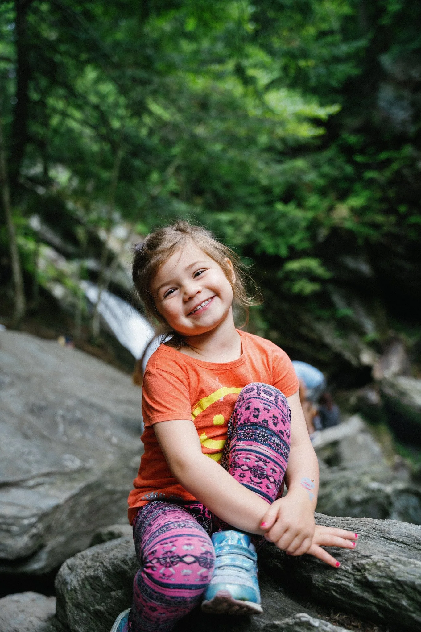A smiling young girl with brown hair, wearing an orange T-shirt and colorful patterned leggings, sitting on a rock in a green forest near a waterfall.