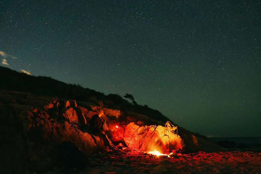 Nighttime beach scene with a campfire, rocky cliff, starry sky, and silhouette of a person sitting near the fire.