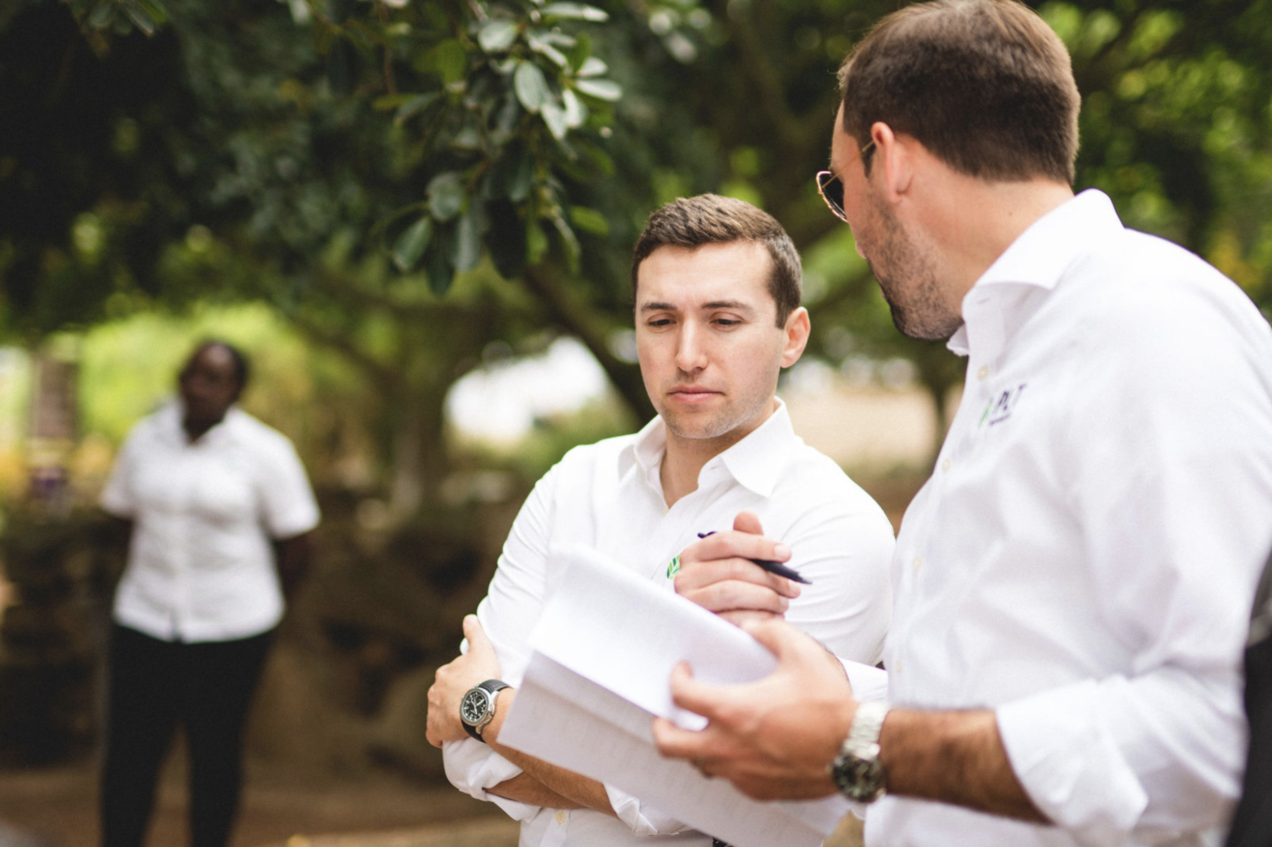 Two men in white shirts discussing documents outdoors with a woman in the background near trees.