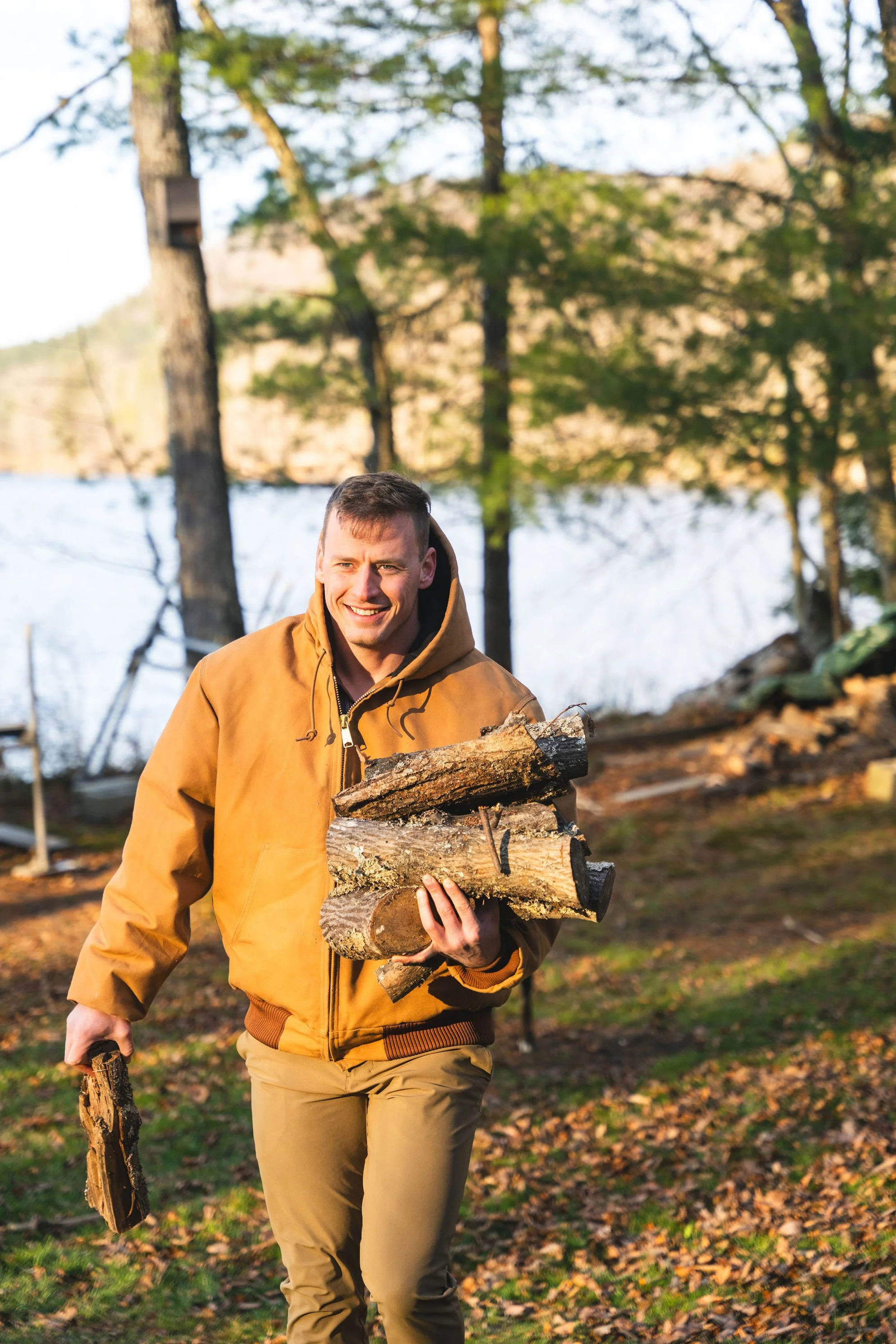A man carrying a stack of firewood outdoors in a forested area near a lake.