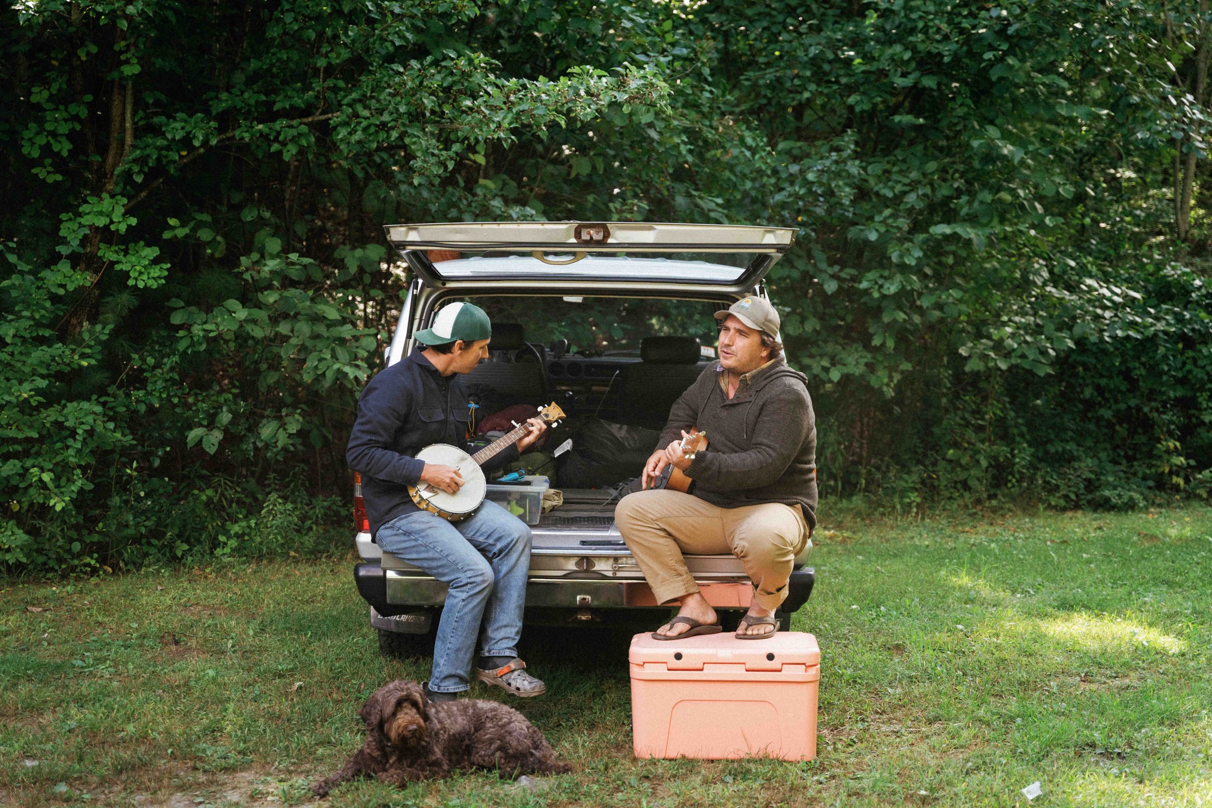 Two men sitting at the back of a car playing guitars and banjo, with a dog lying on the grass nearby.