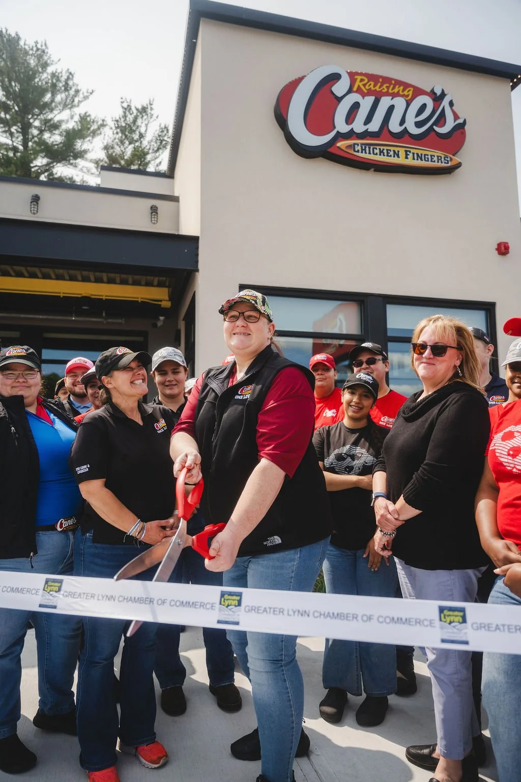 Group of people gathered outside a Raising Cane's restaurant during a ribbon-cutting event, with one person holding scissors.