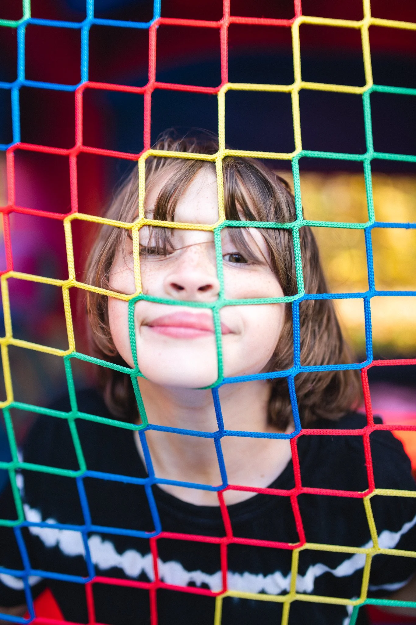 A child with shoulder-length brown hair behind a colorful playground net, smiling at the camera.