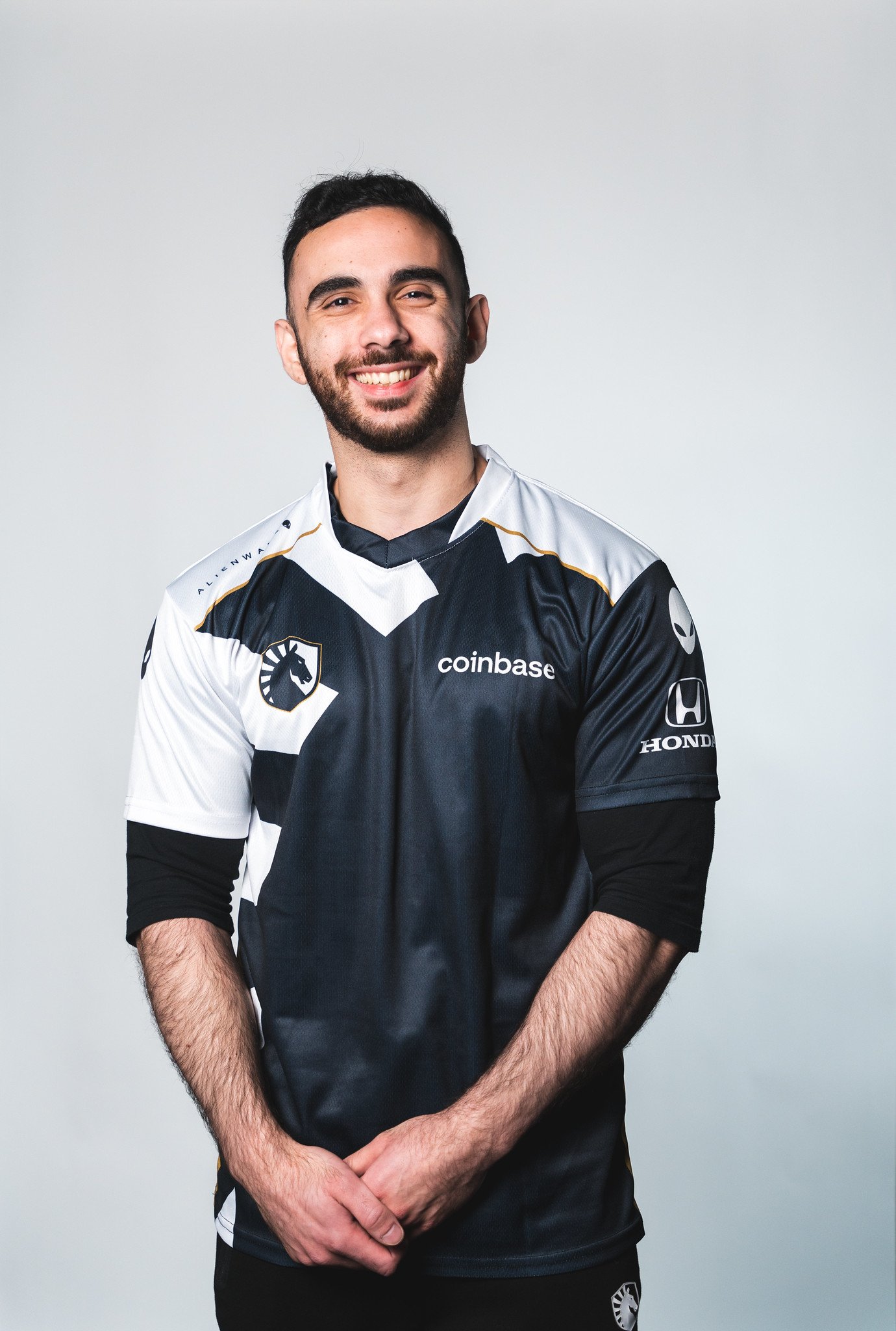 A young man with dark hair and a beard smiling, wearing a black and white esports jersey with team and sponsor logos, standing against a plain light gray background.