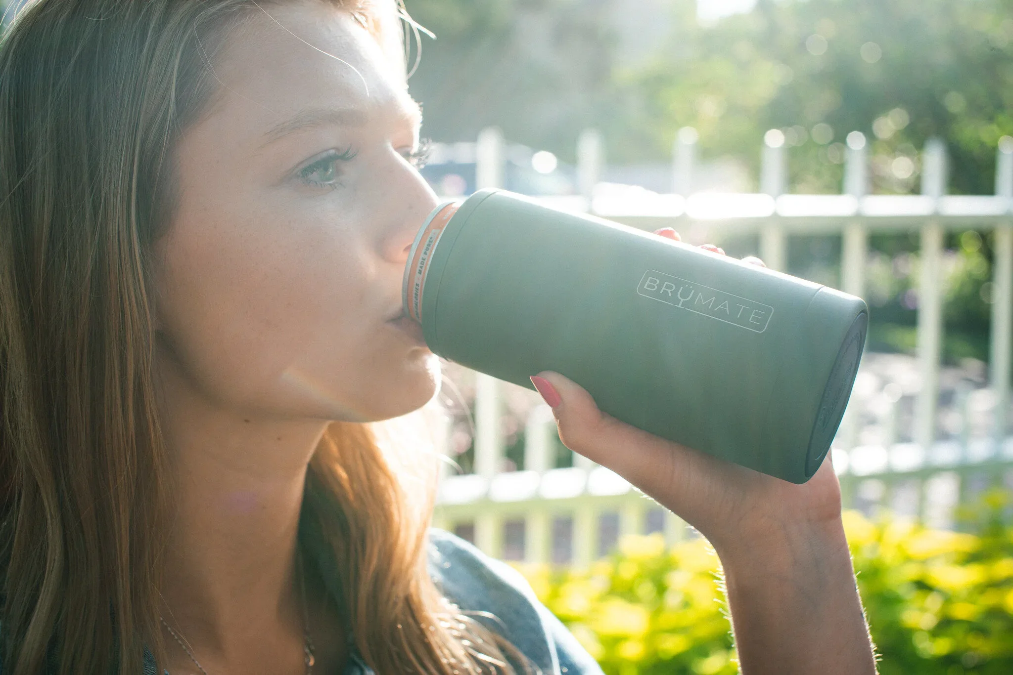 A young woman with light skin and red hair drinks from a green Brumate tumbler outdoors on a sunny day, with greenery and a white fence in the background.