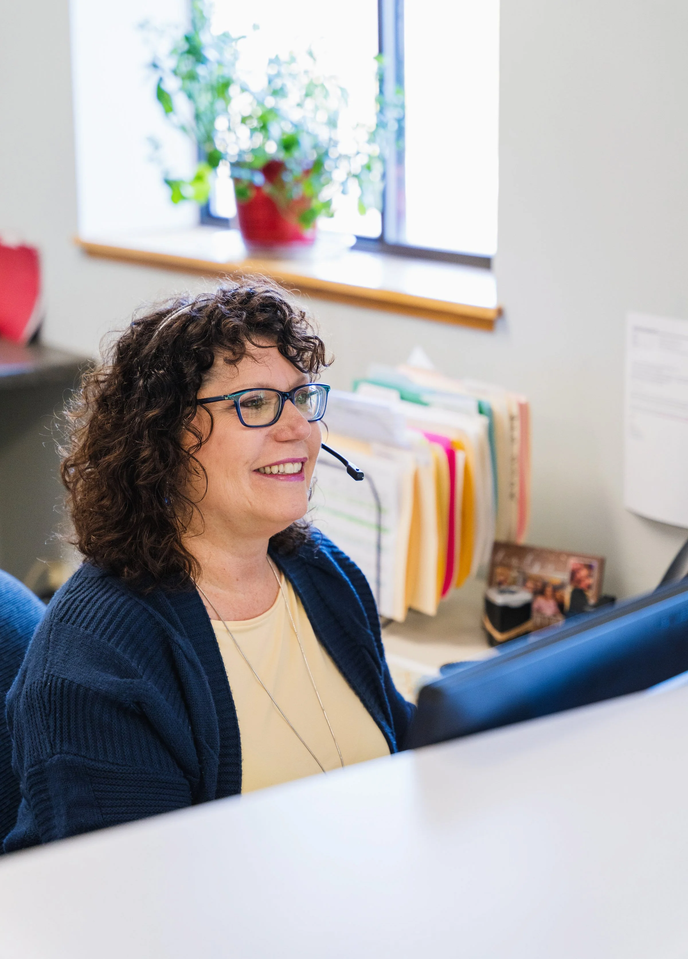 A woman with curly brown hair, glasses, and a headset smiling at her desk in an office.