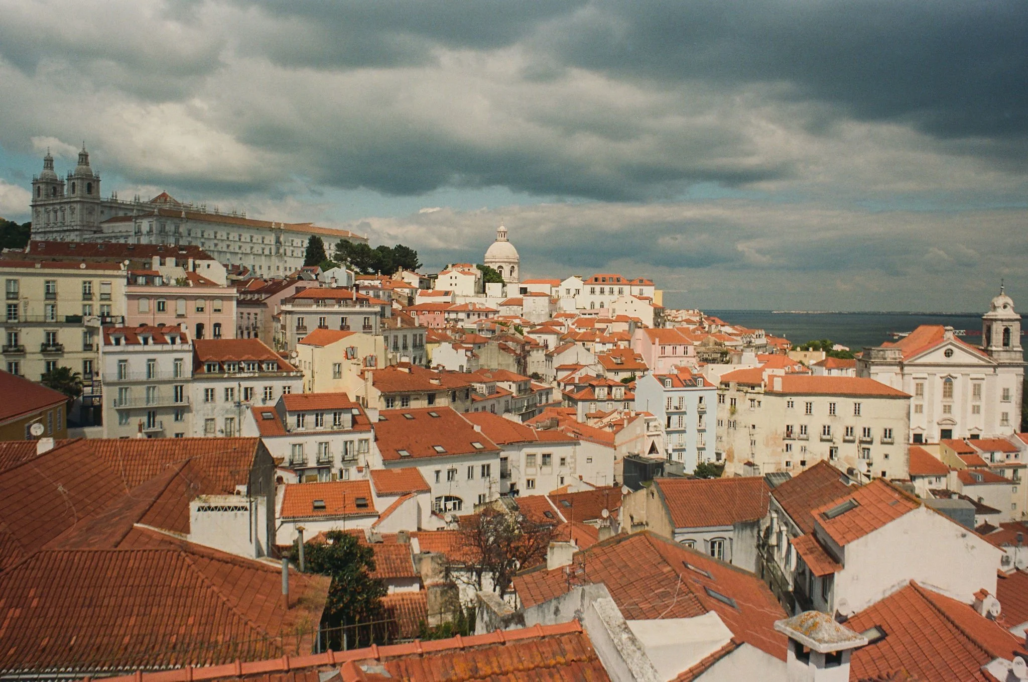 A cityscape with a dense cluster of buildings with red-tiled roofs, a large historic cathedral at the hilltop, and a body of water in the distance under a cloudy sky.