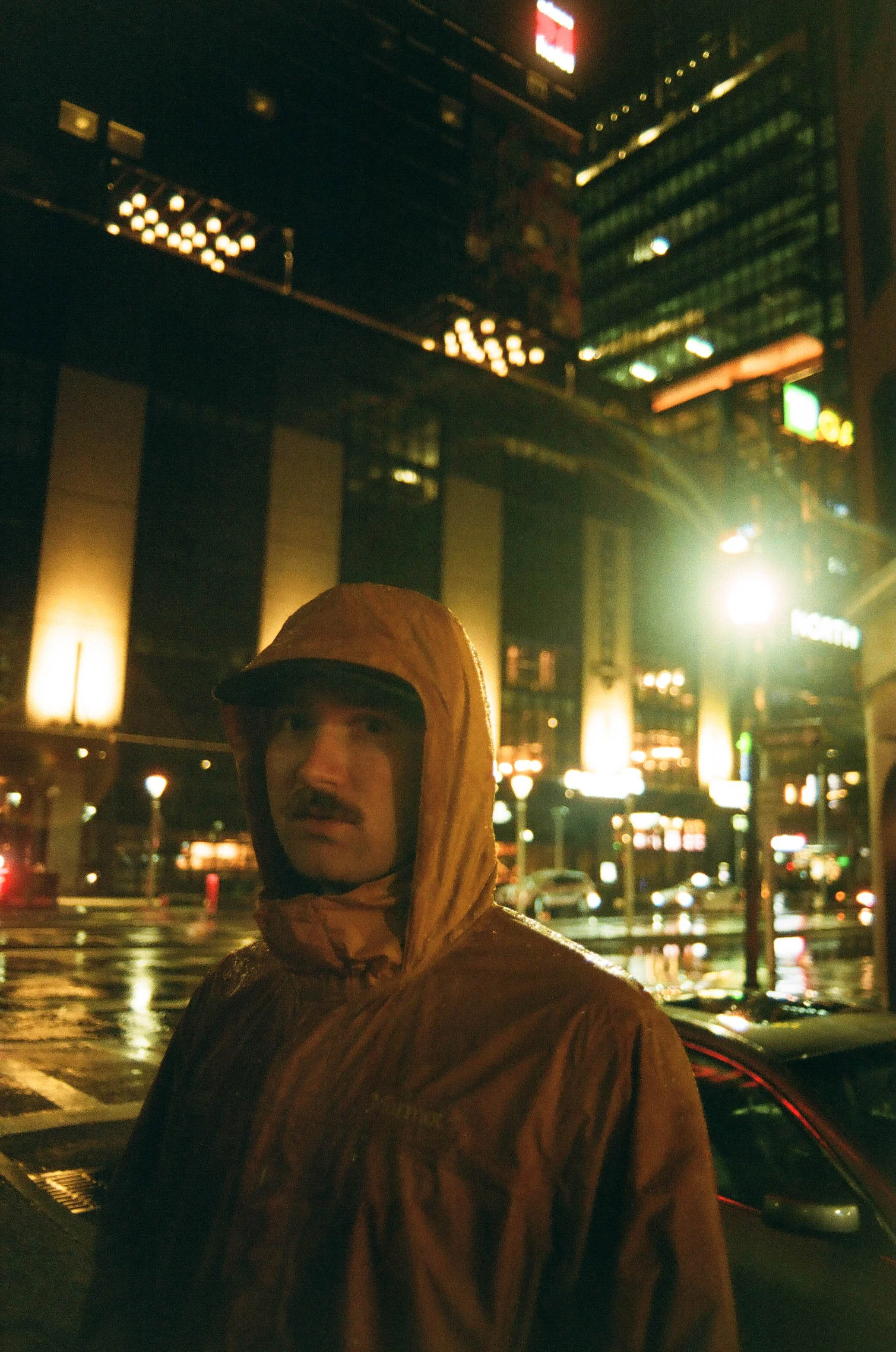 A man wearing a brown raincoat with a hood on a rainy night in an urban setting, illuminated by city lights and reflections on wet streets.