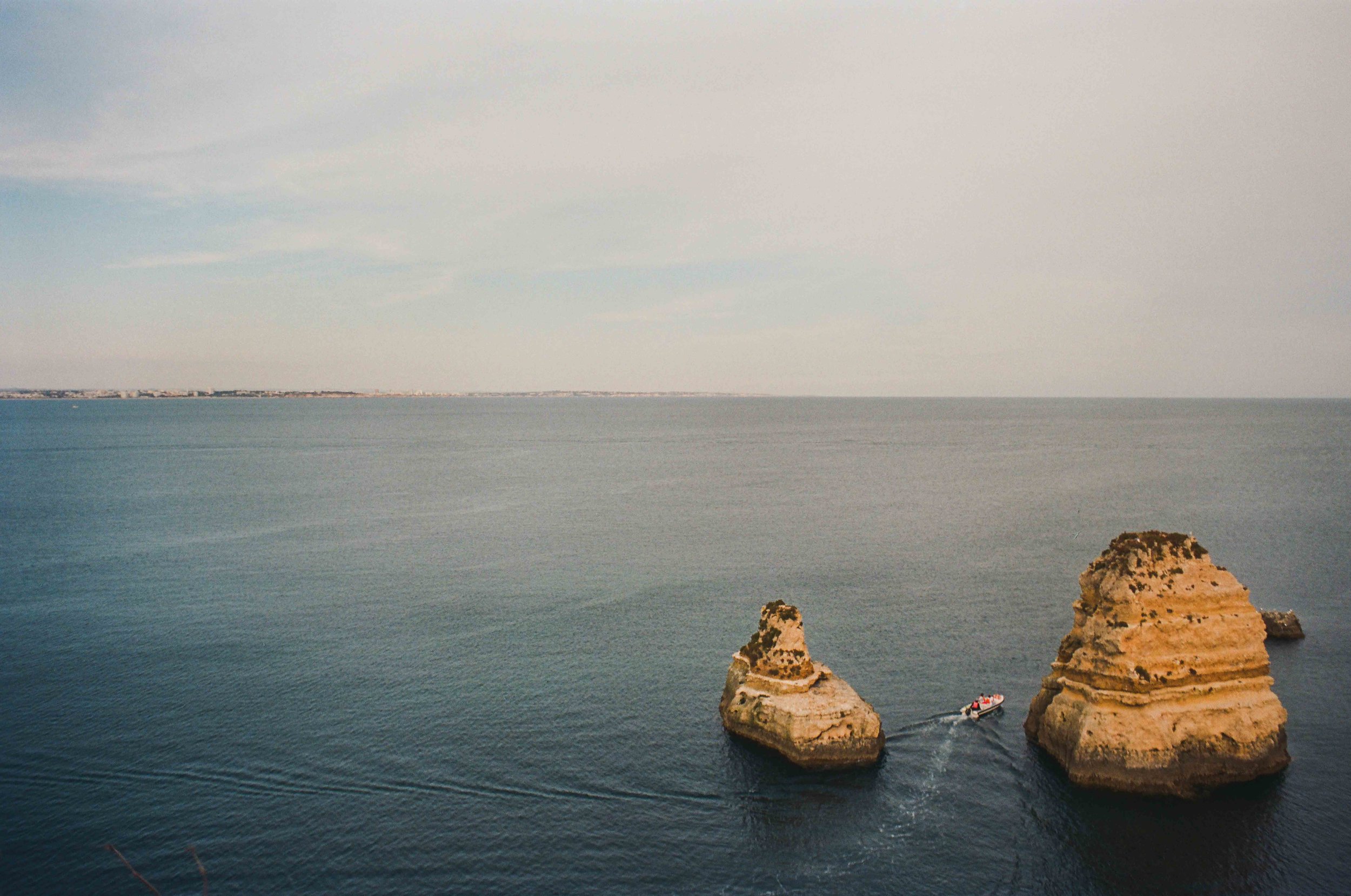 Two large rock formations in the ocean with a boat sailing between them, and a distant shoreline on the horizon.
