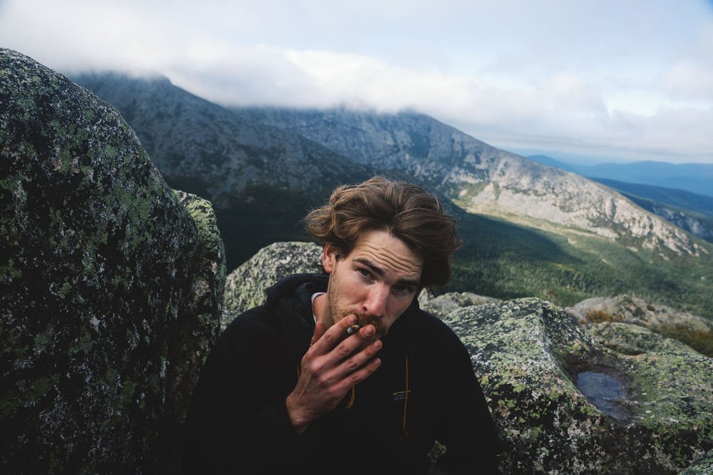 A young man with tousled brown hair, wearing a black jacket, is sitting on rocks in a mountainous landscape with a cloudy sky in the background.