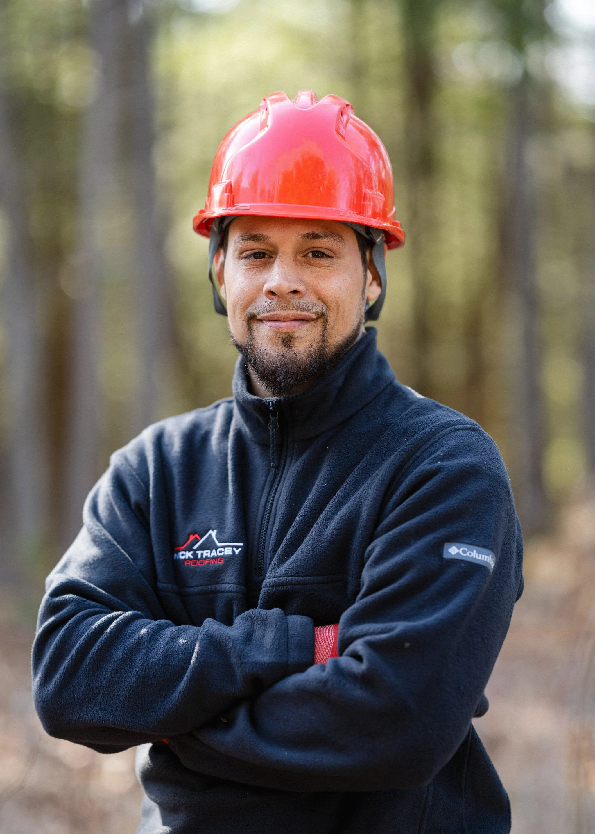 A man wearing a red safety helmet and black Columbia fleece jacket crosses his arms outdoors in a wooded area, smiling slightly.