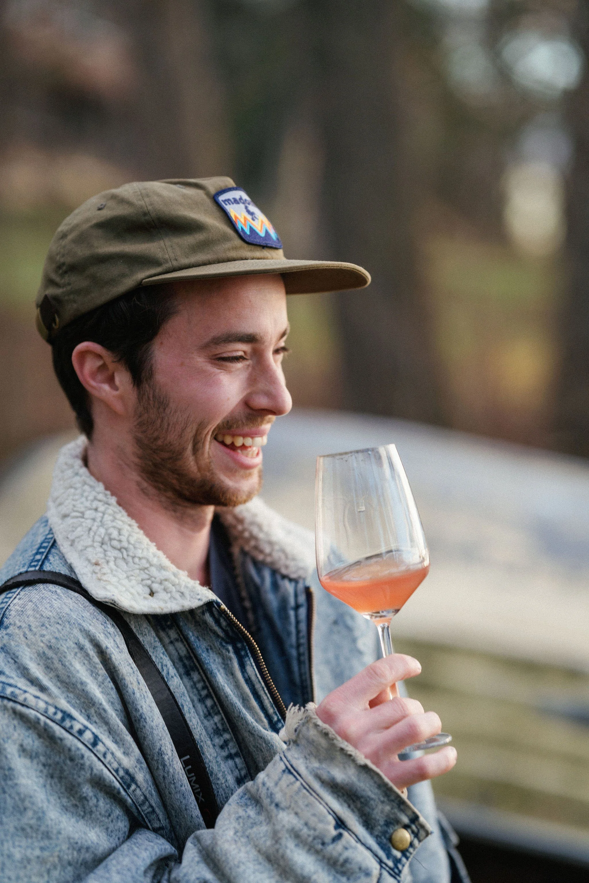 A young man with a beard smiling and looking at a glass of rosé wine outdoors during daytime, wearing a tan cap, a denim jacket with a shearling collar, and a black strap over his shoulder.