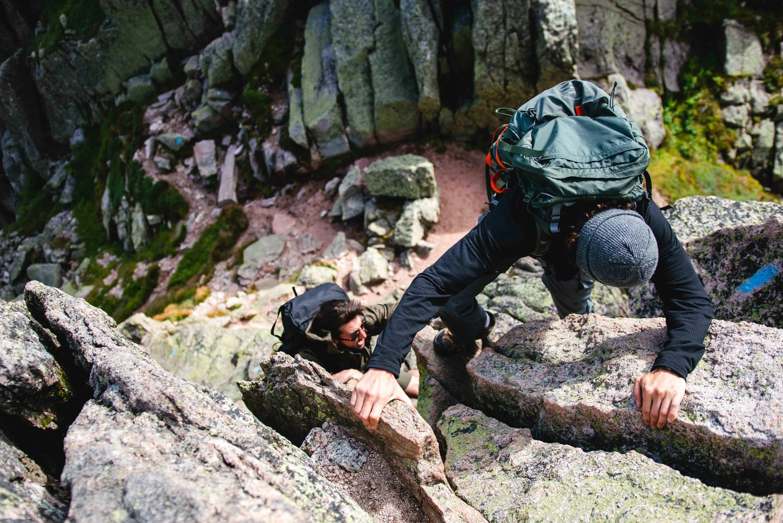 Two hikers climbing a rocky trail on Mount Katahdin, both carrying backpacks and dressed in outdoor gear.