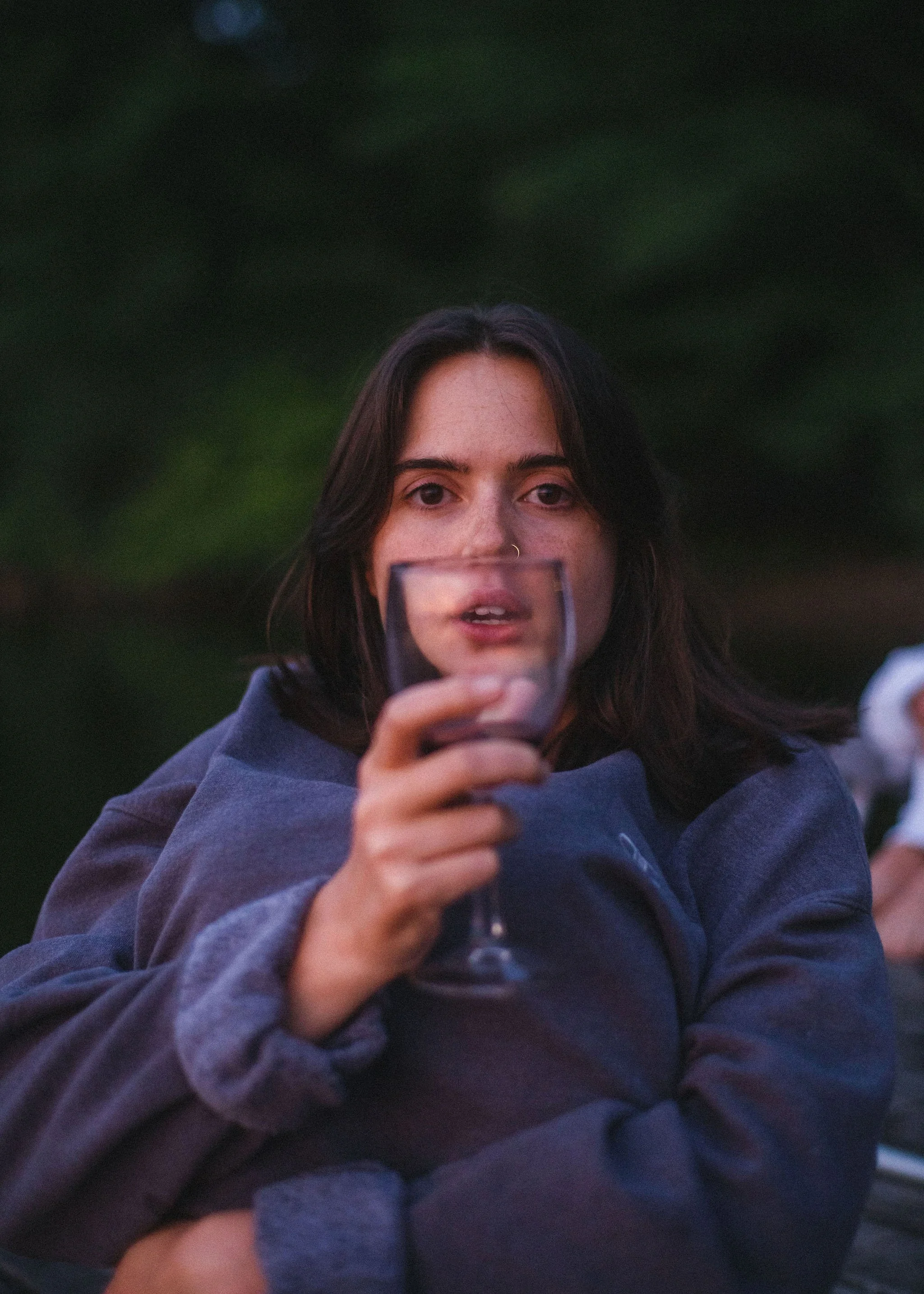 A woman with dark hair, wearing a dark jacket, holding a glass of red wine, sitting outdoors with a blurred green background.