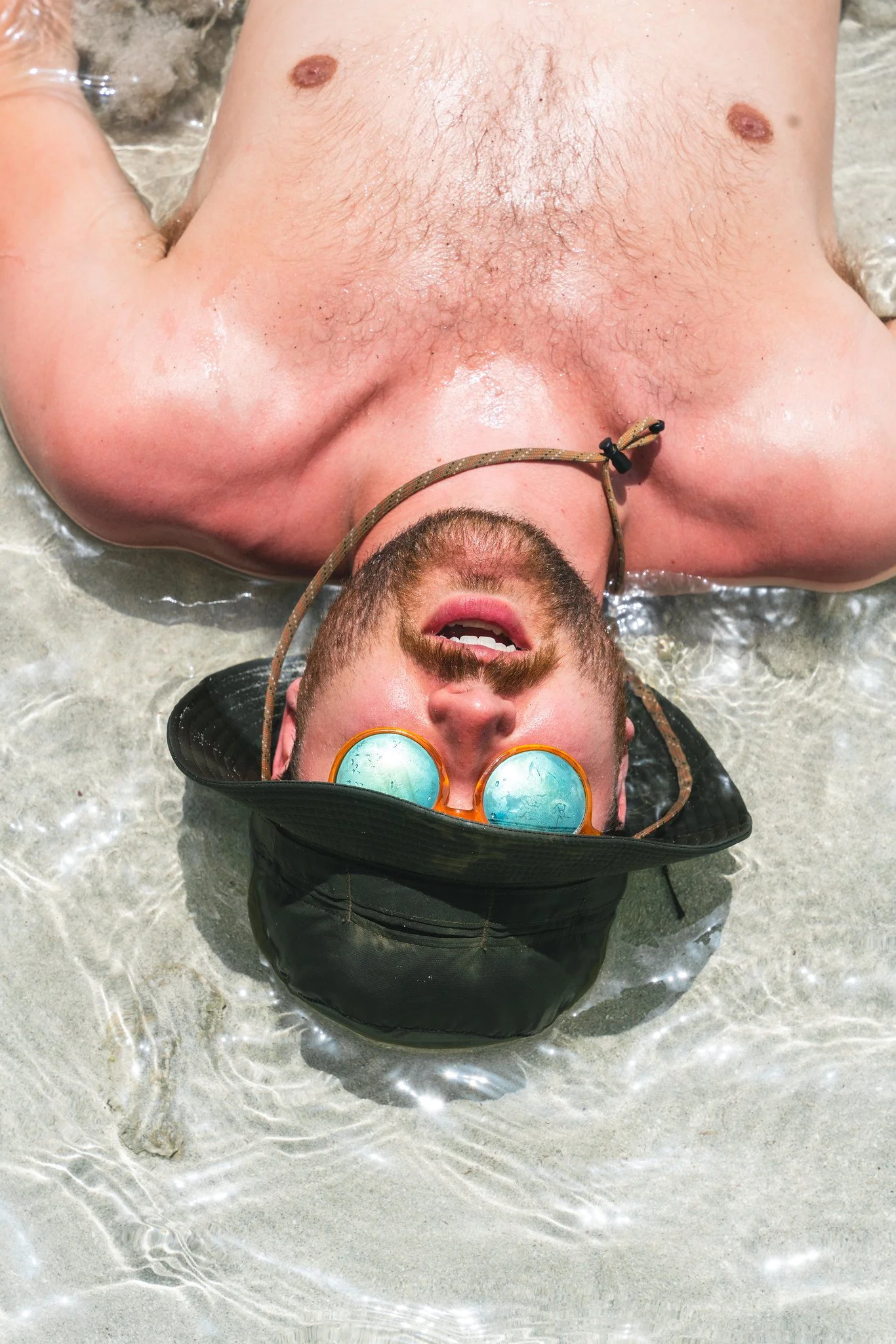 A man with a beard and sunglasses lying on his back in shallow water on the beach, wearing a black hat and a necklace.