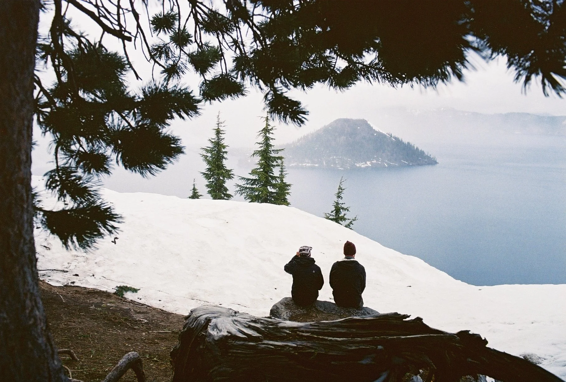 Two people sitting on a rock overlooking a snowy landscape with evergreen trees, a body of water, and a distant mountain.