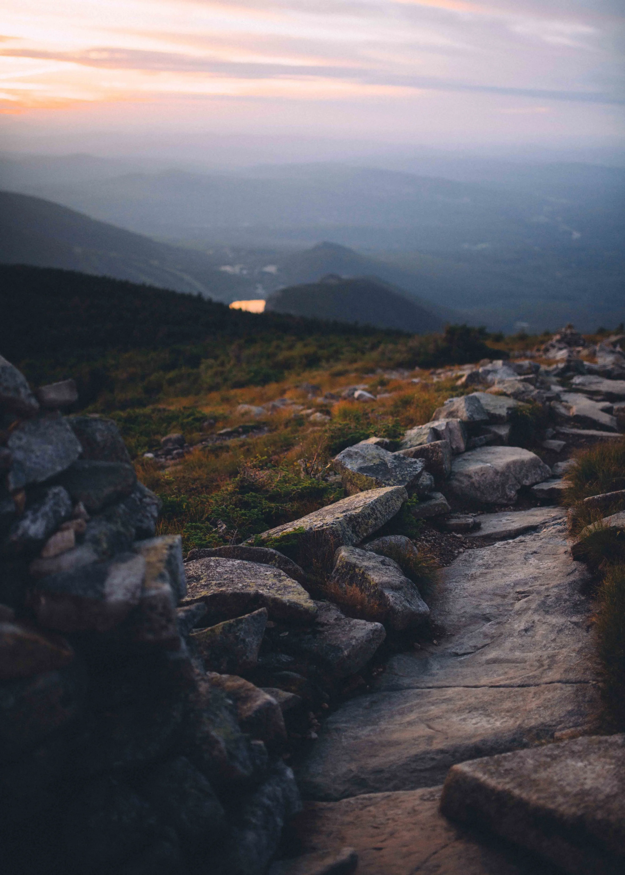 A rugged mountain trail with rocks and greenery, overlooking a series of mountain ridges under a sunset sky.