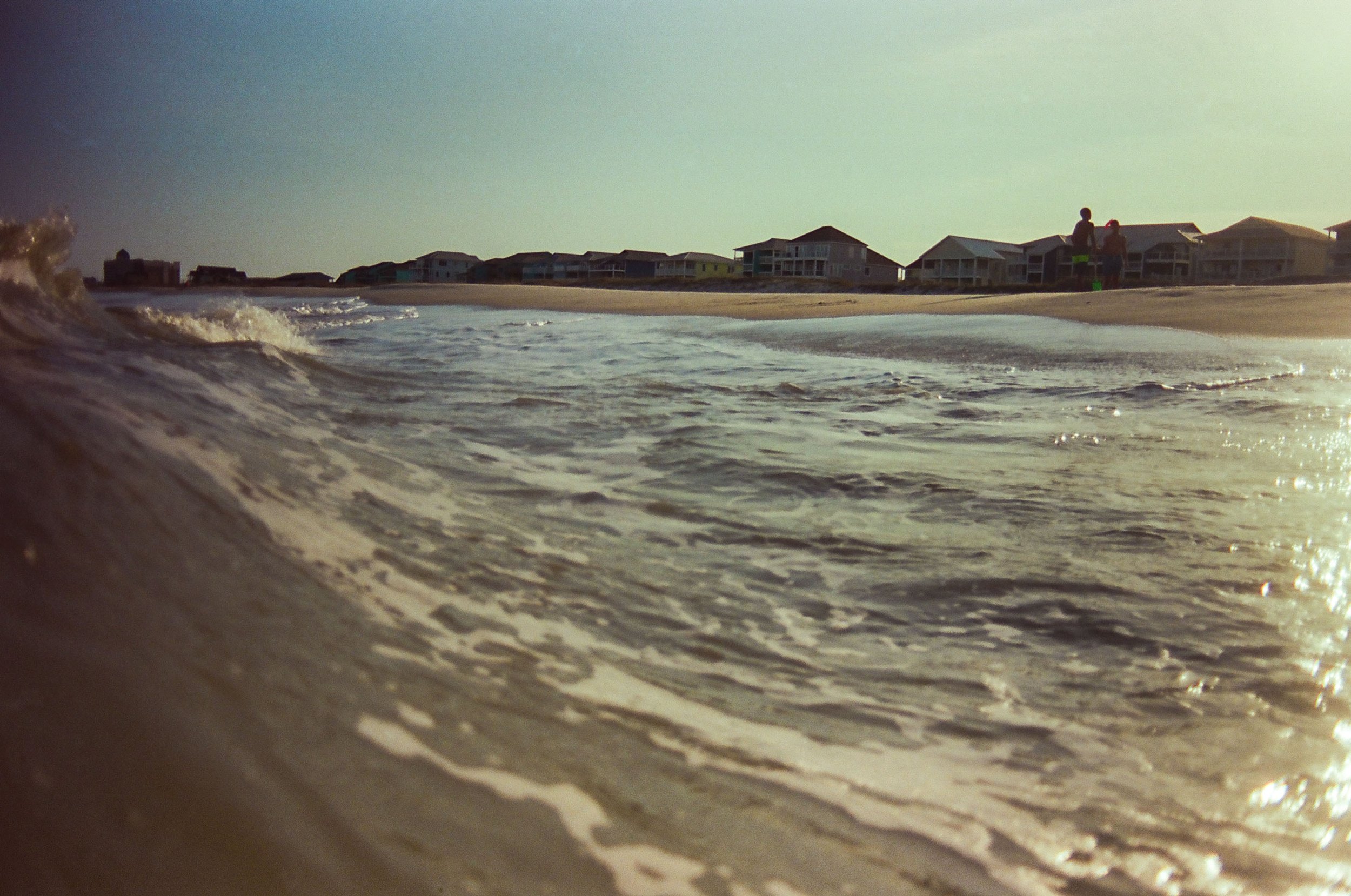 Beach scene with gentle waves washing onto shore, houses visible in the distance, and two people standing on the beach.