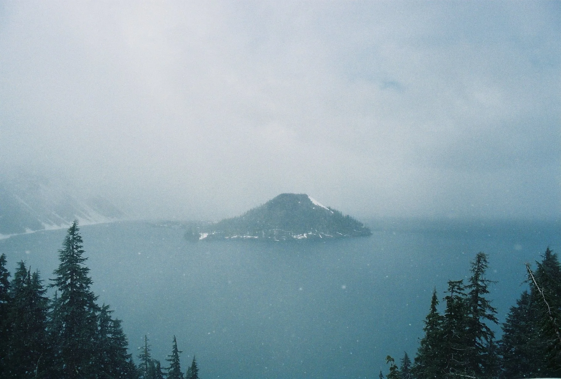 A large body of water with an island in the middle, surrounded by dense evergreen trees, and misty mountains in the background on a cloudy, foggy day.
