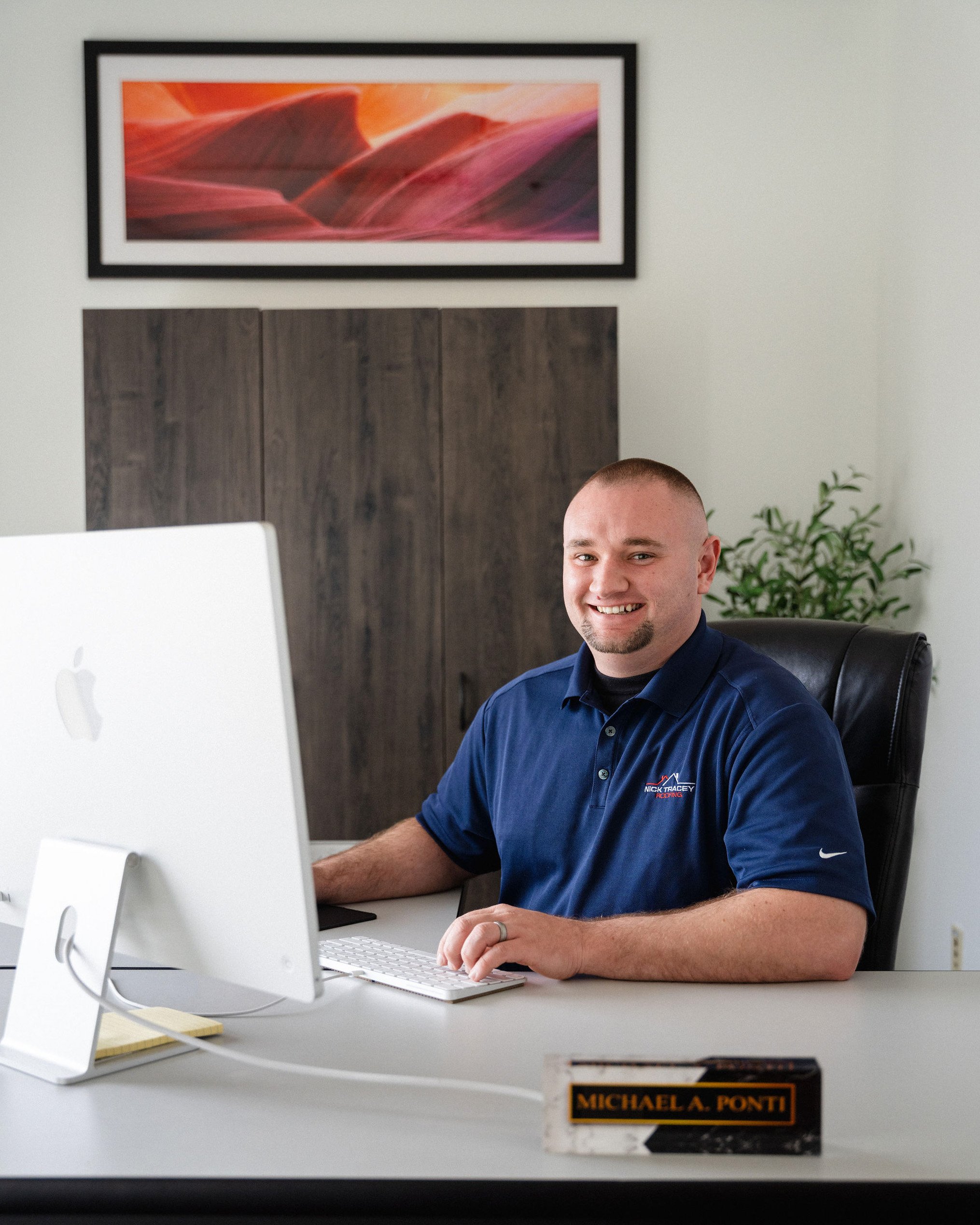 A man sitting at a desk with an Apple iMac, smiling, wearing a navy blue polo shirt, in an office with a framed abstract art piece and a potted plant in the background.