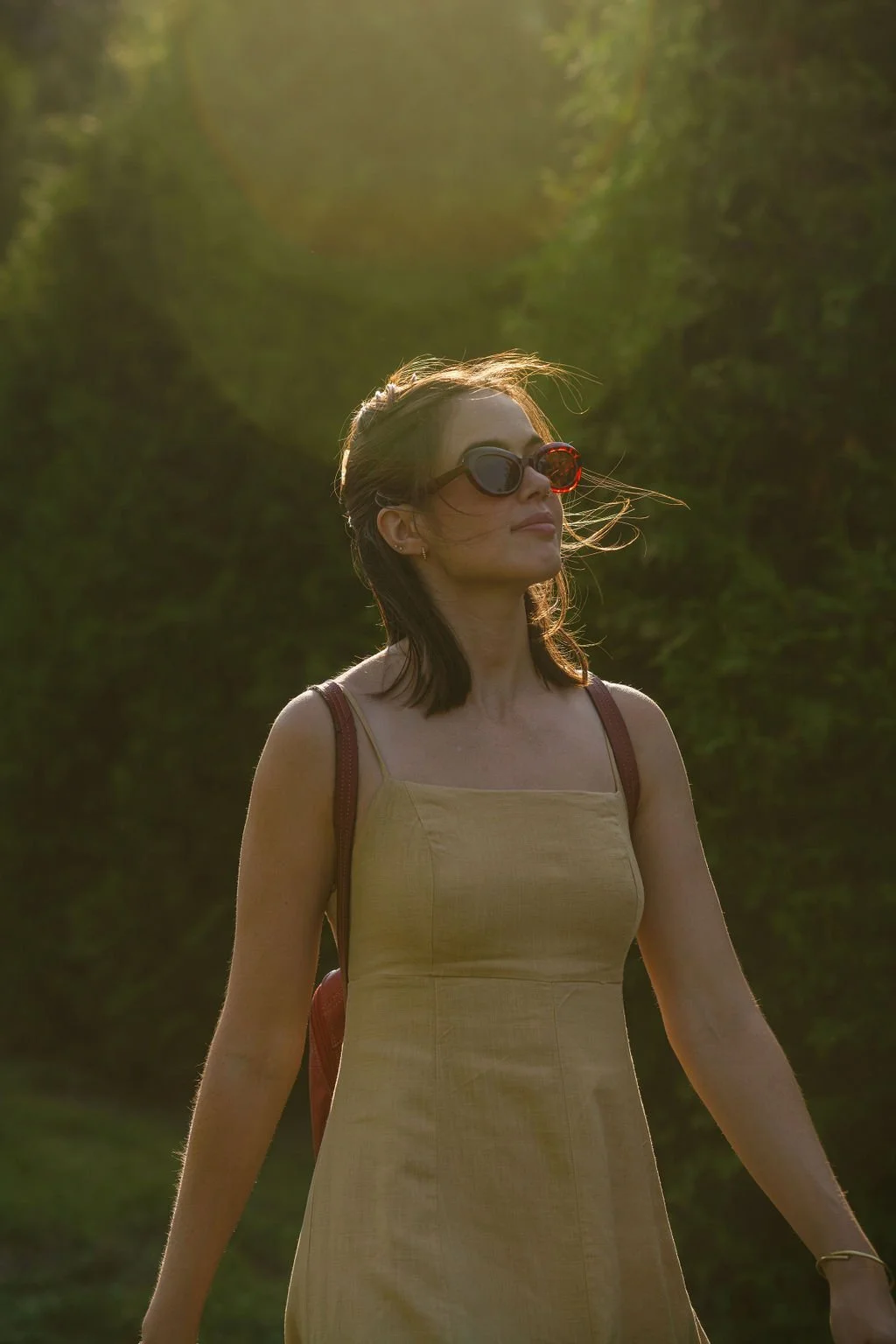 A woman wearing sunglasses and a yellow dress walking outdoors in natural sunlight, with greenery in the background.