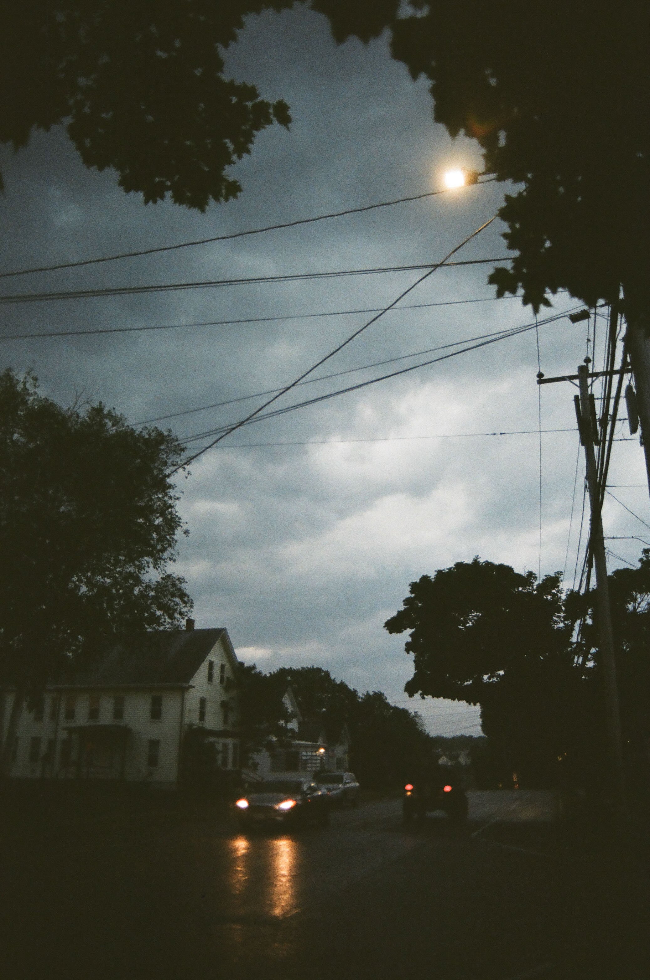 Nighttime street scene with cars, houses, trees, and a cloudy sky with the moon visible.