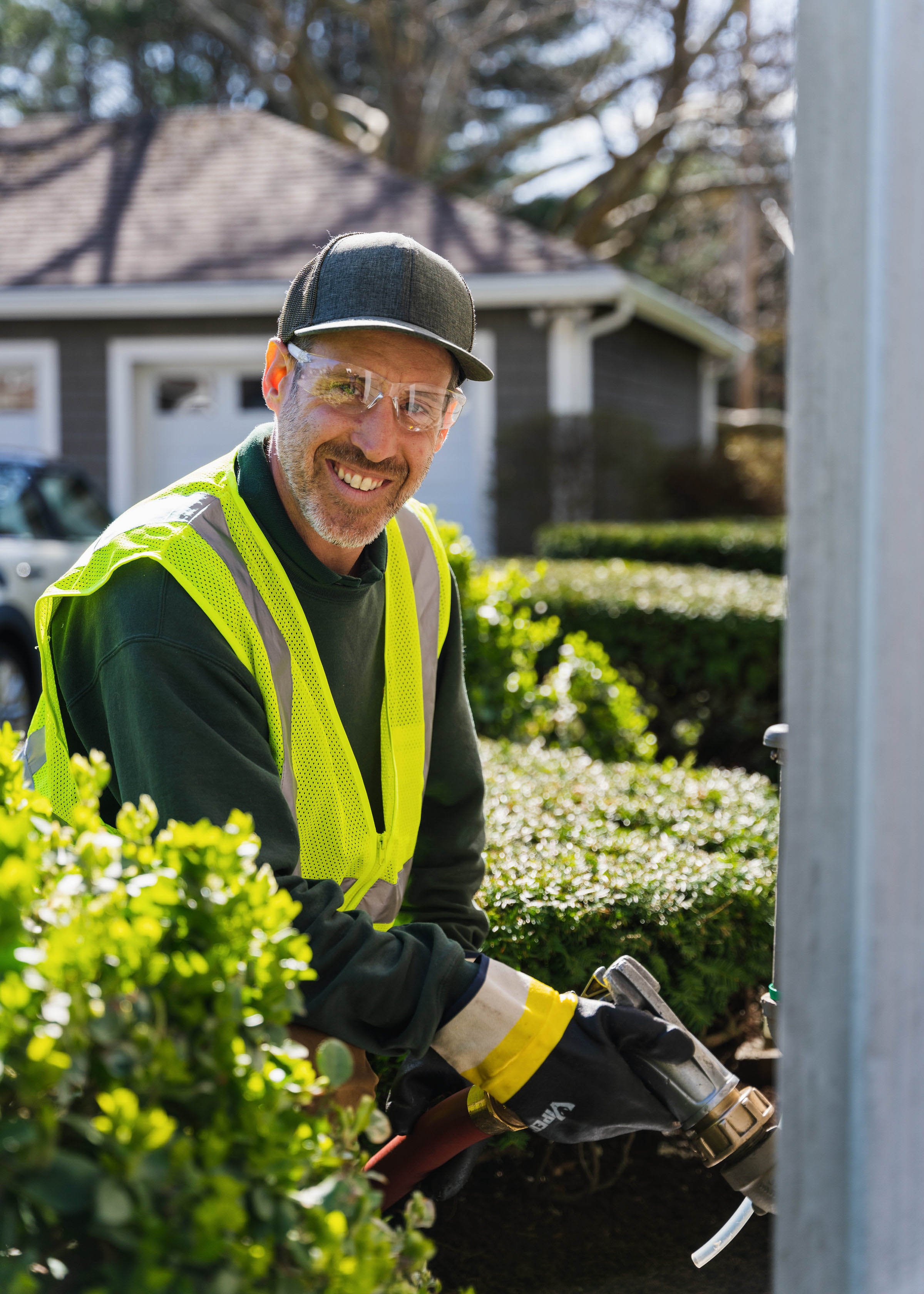 A smiling man wearing safety glasses, a black cap, a yellow safety vest, and gloves, working outdoors with gardening equipment near bushes and a house.