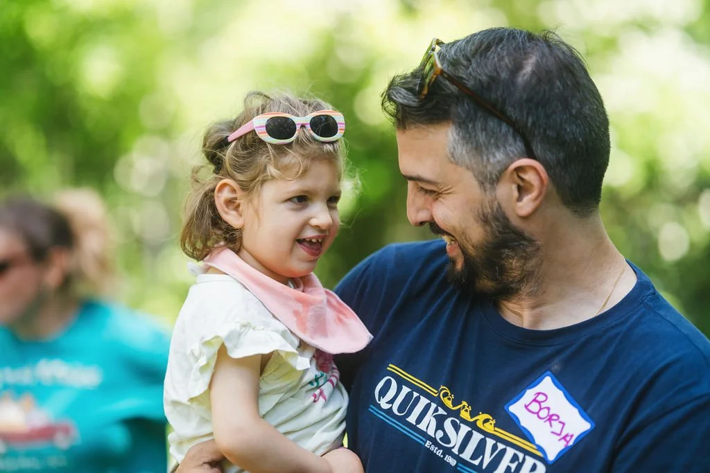 A man holding a young girl outdoors, both smiling and looking at each other, with a background of green trees.