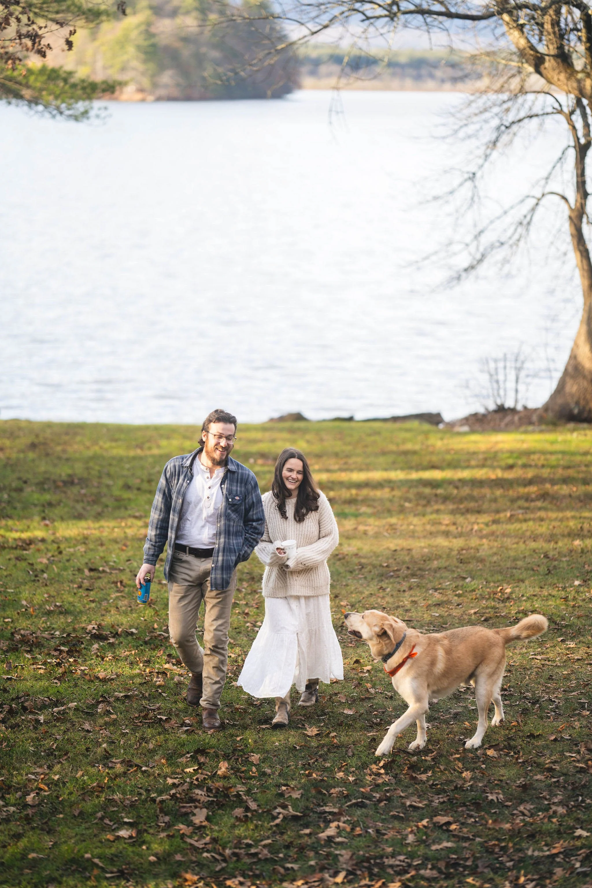 A happy couple walking with their dog near a lake on a fall day, smiling and enjoying each other's company.
