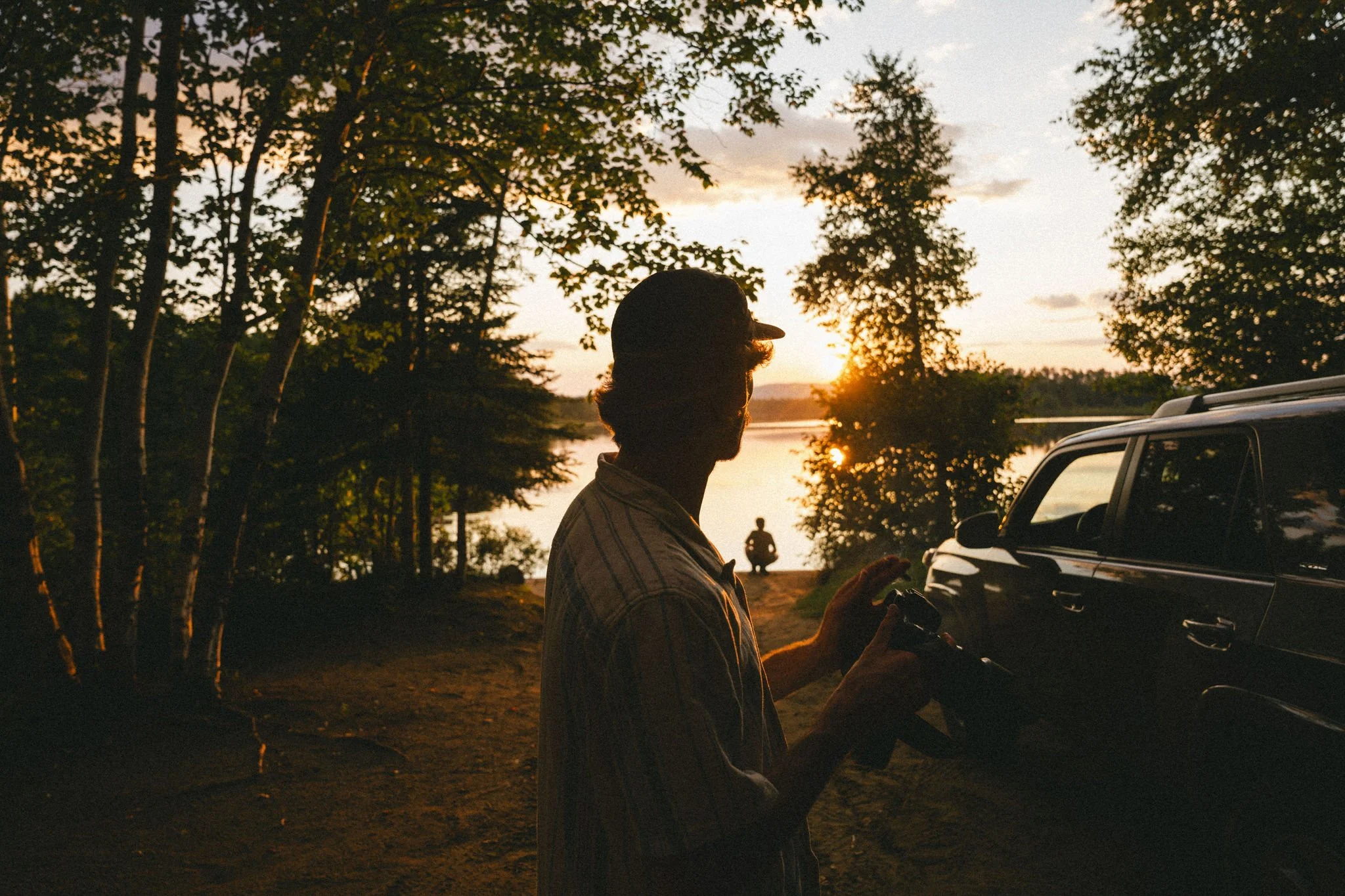 A person holding a camera standing next to a vehicle outdoors during sunset, with a lake and trees in the background and another person sitting by the water.