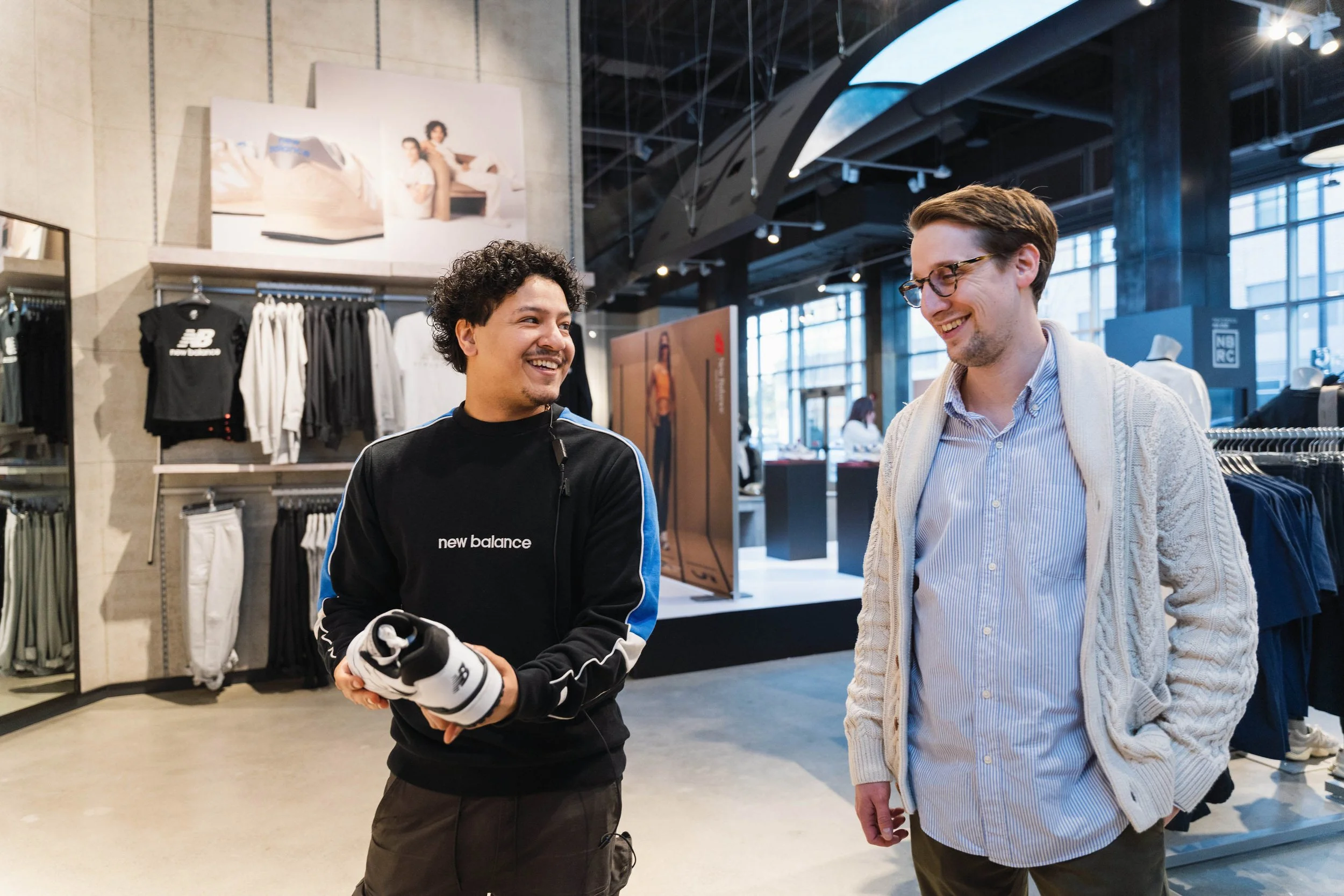 Two men smiling and talking inside a retail clothing store, surrounded by racks of clothing and posters on the walls.