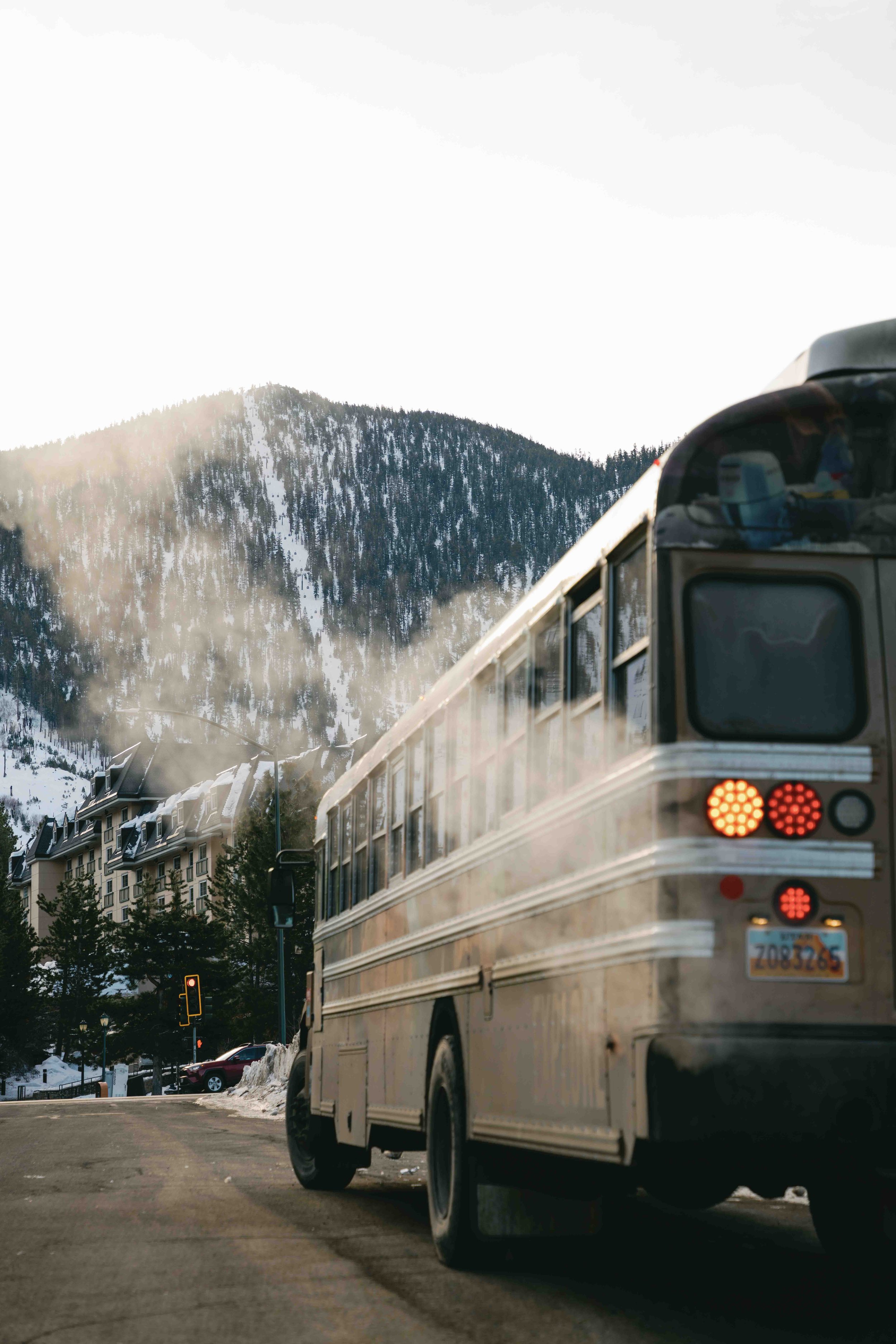 A city bus on a mountain road with snow-covered mountains in the background and buildings on the side.