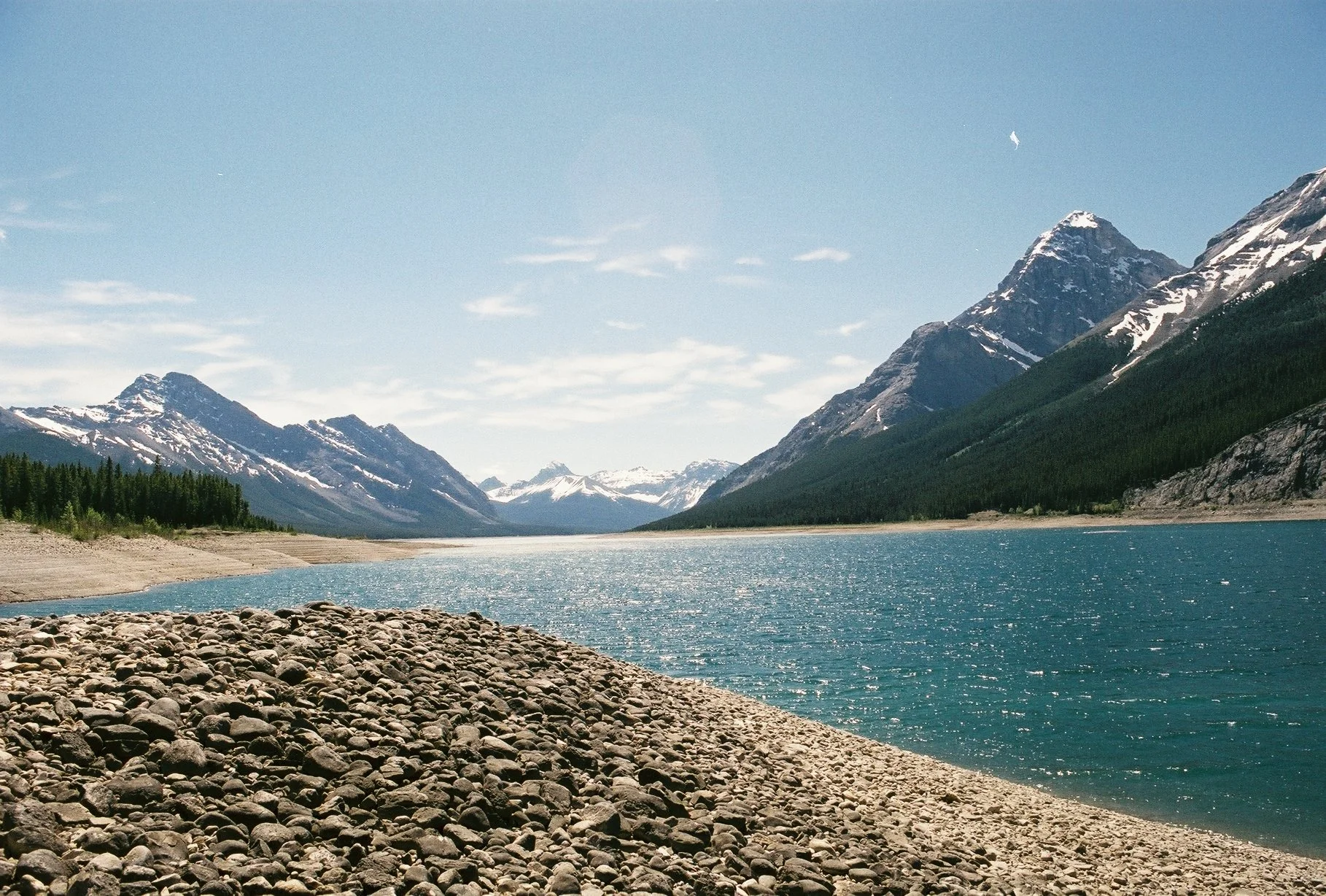 A lake surrounded by snow-capped mountains and green forests, with a rocky shoreline in the foreground under a clear blue sky.