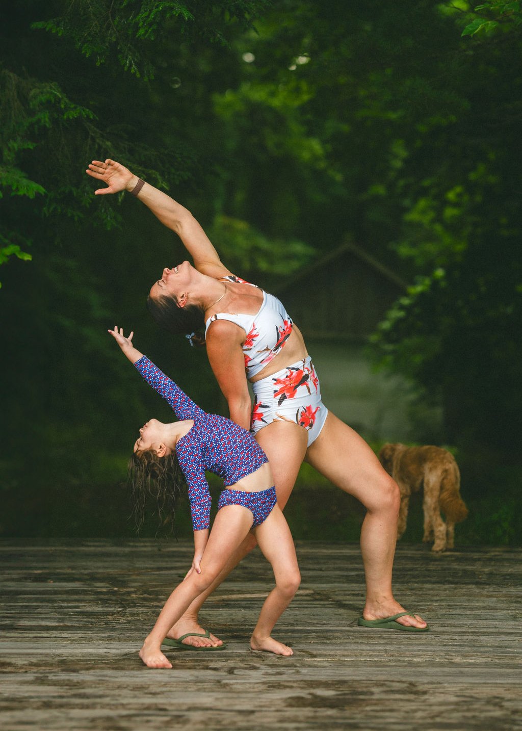 Two women practicing yoga outdoors on a wooden deck with lush green trees in the background. One woman, in a floral two-piece outfit, is standing and arching back with her arm extended upward. The other woman, in a patterned outfit, is bent forward w