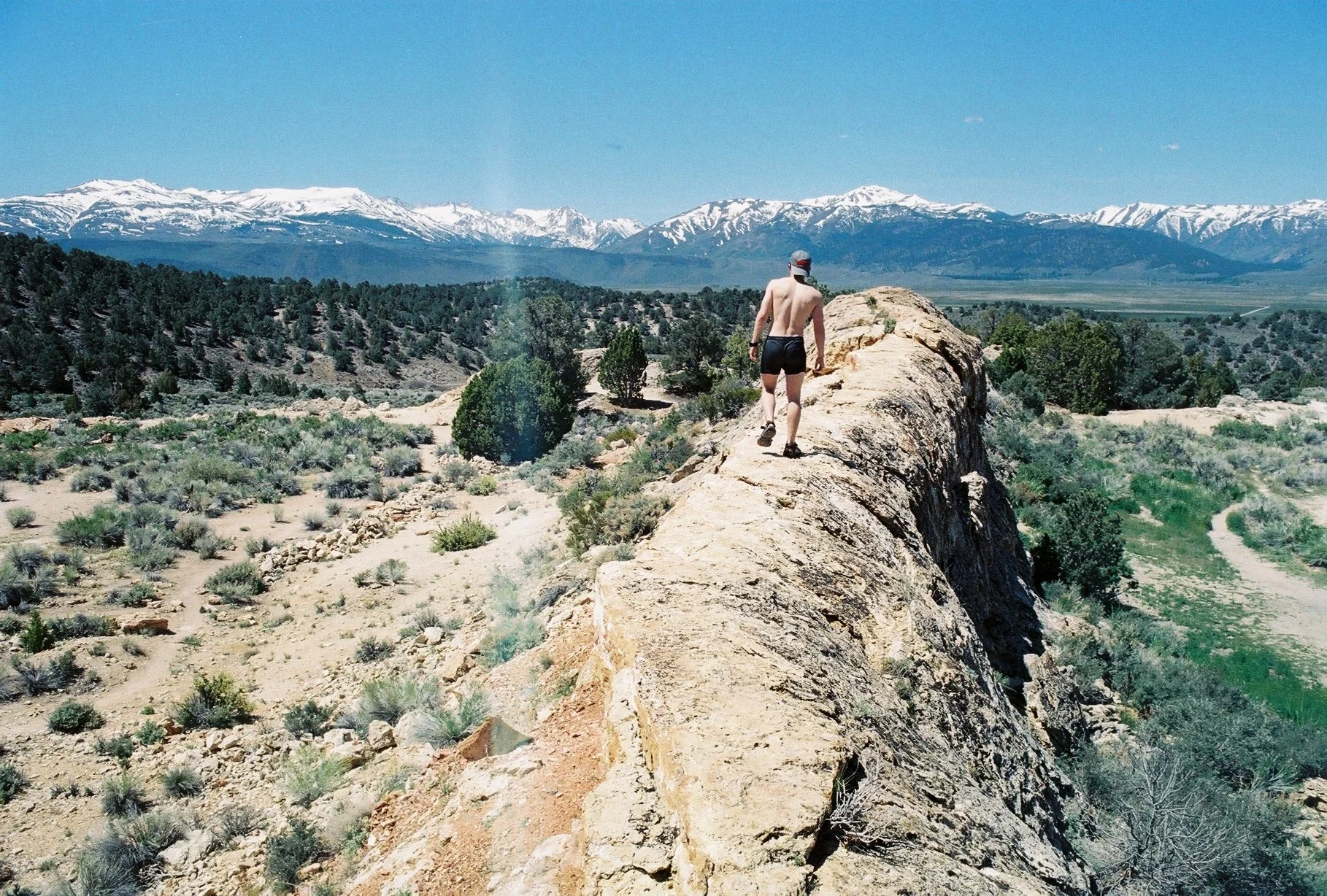 A man in black shorts, a hat, and sunglasses walking along a rocky ridge in a desert landscape, with snow-capped mountains in the distance, under a clear blue sky.