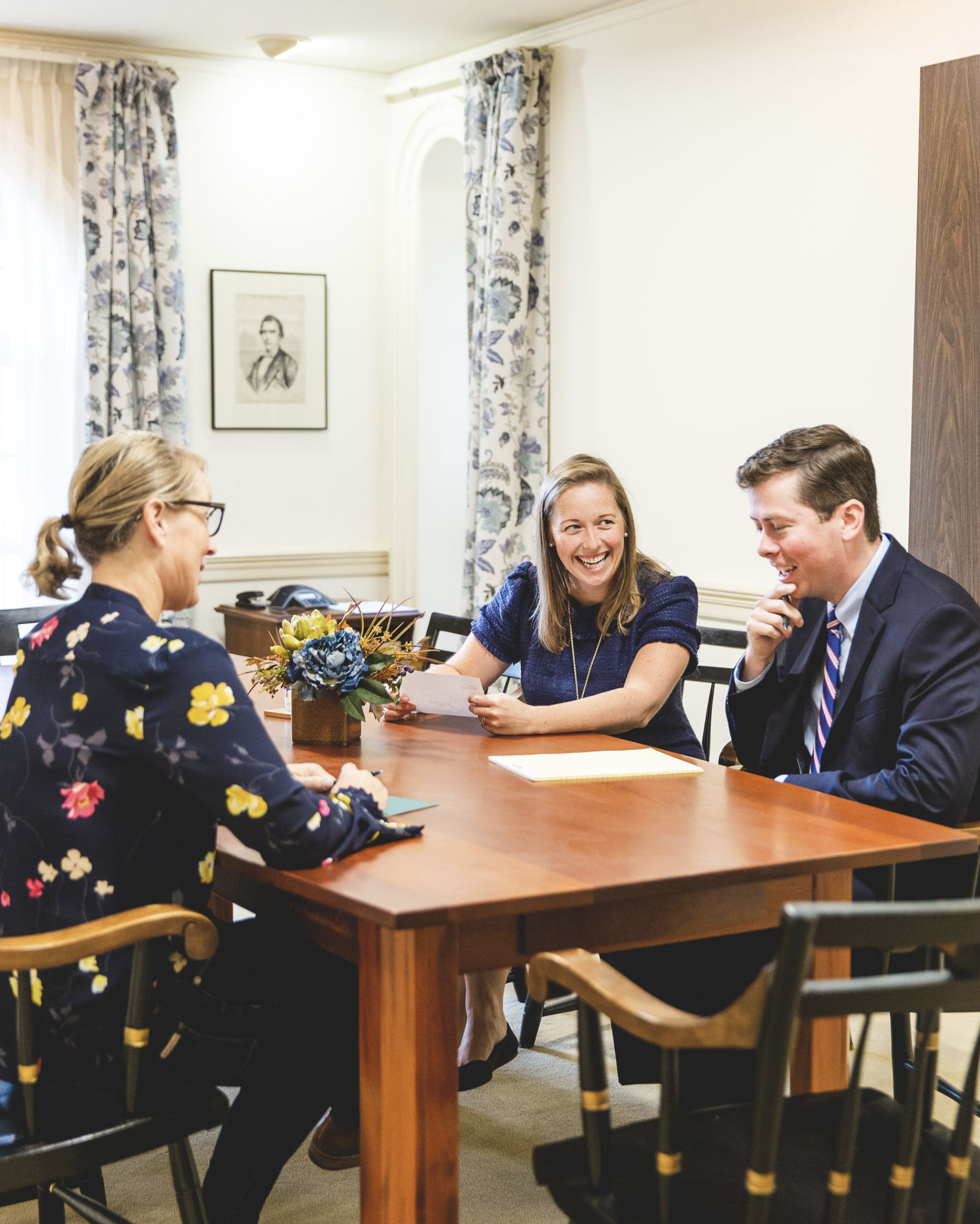 Three people sitting at a wooden table in a meeting room, engaged in conversation and smiling, with a woman holding papers, a man in a suit, and another woman with glasses and a floral blouse.