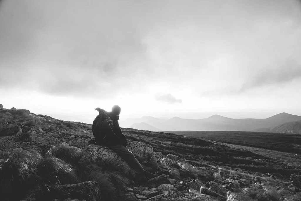 A person sitting on rocks in a mountainous landscape, looking toward the horizon with mountains in the distance under a cloudy sky.