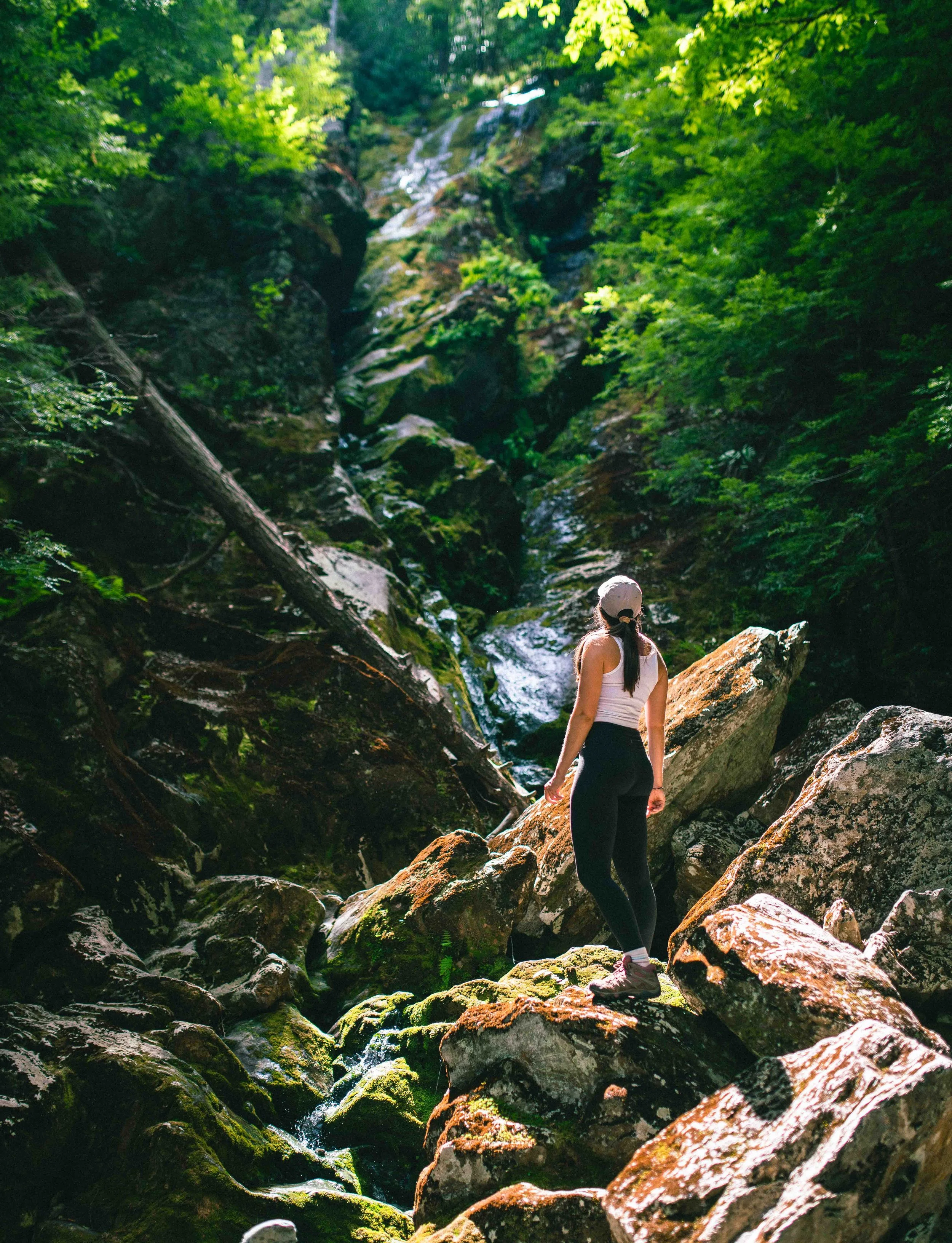 A woman standing on moss-covered rocks near a small waterfall in a lush green forest.