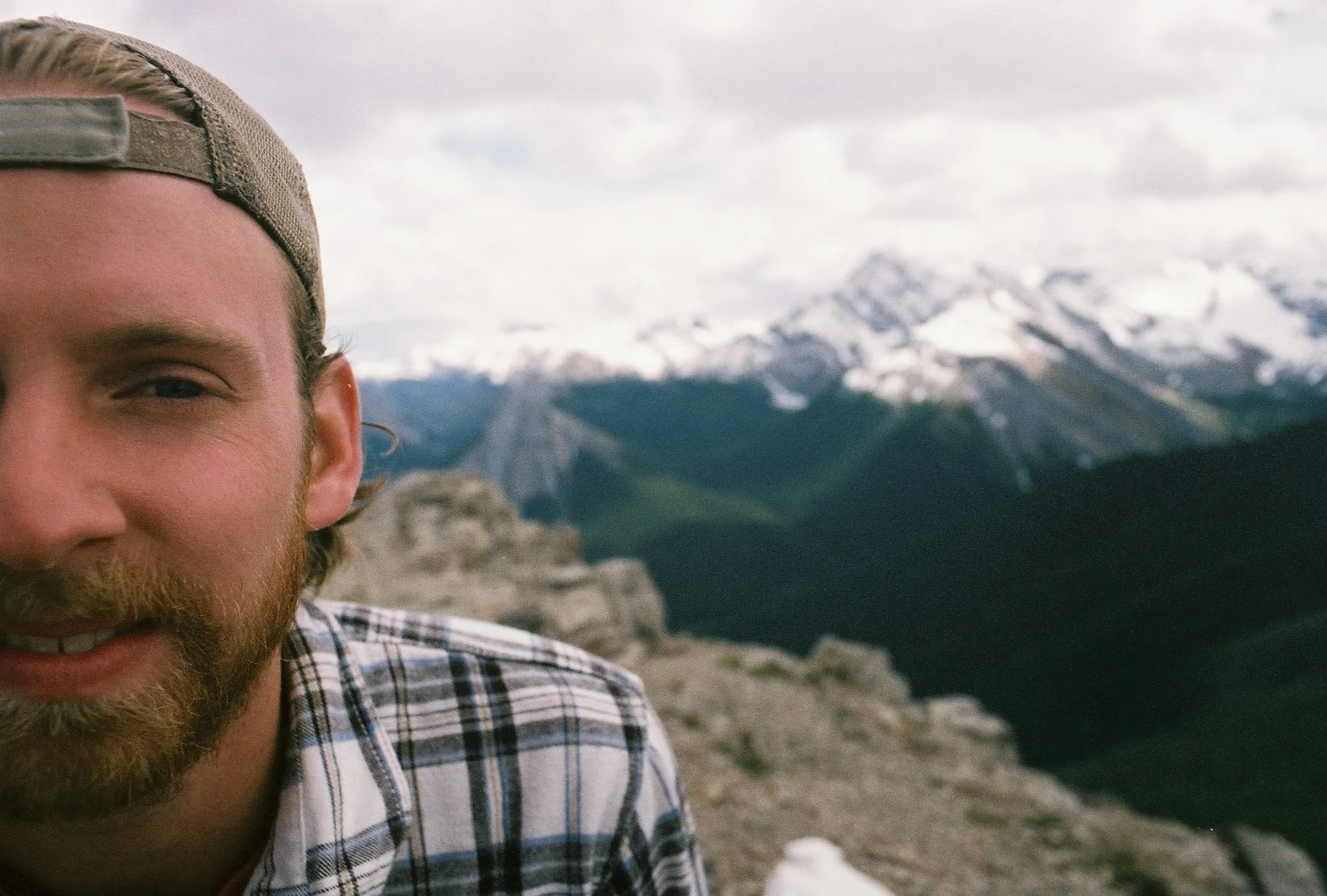 Partial view of a man with a beard and baseball cap, smiling in front of snow-capped mountains.