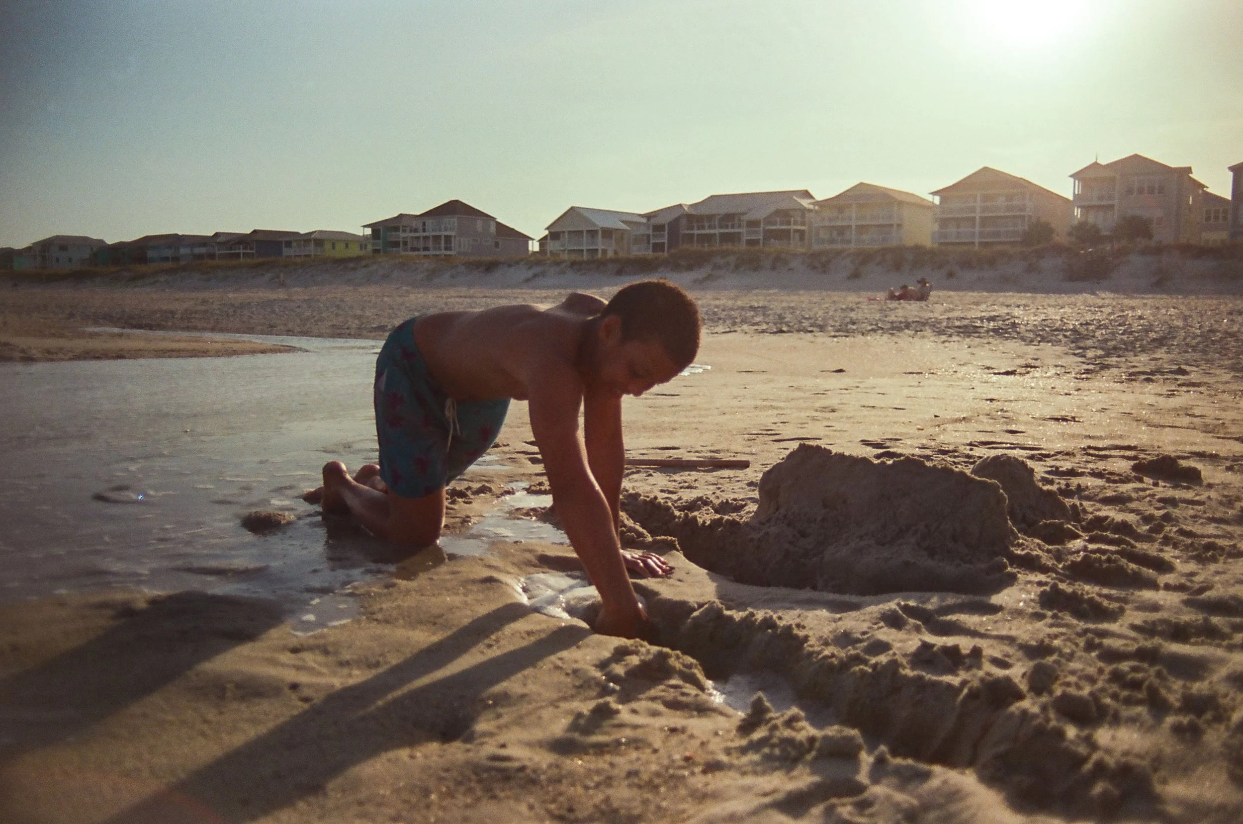 A young boy kneeling in shallow water on a sandy beach, digging in the sand during sunset, with beach houses in the background.