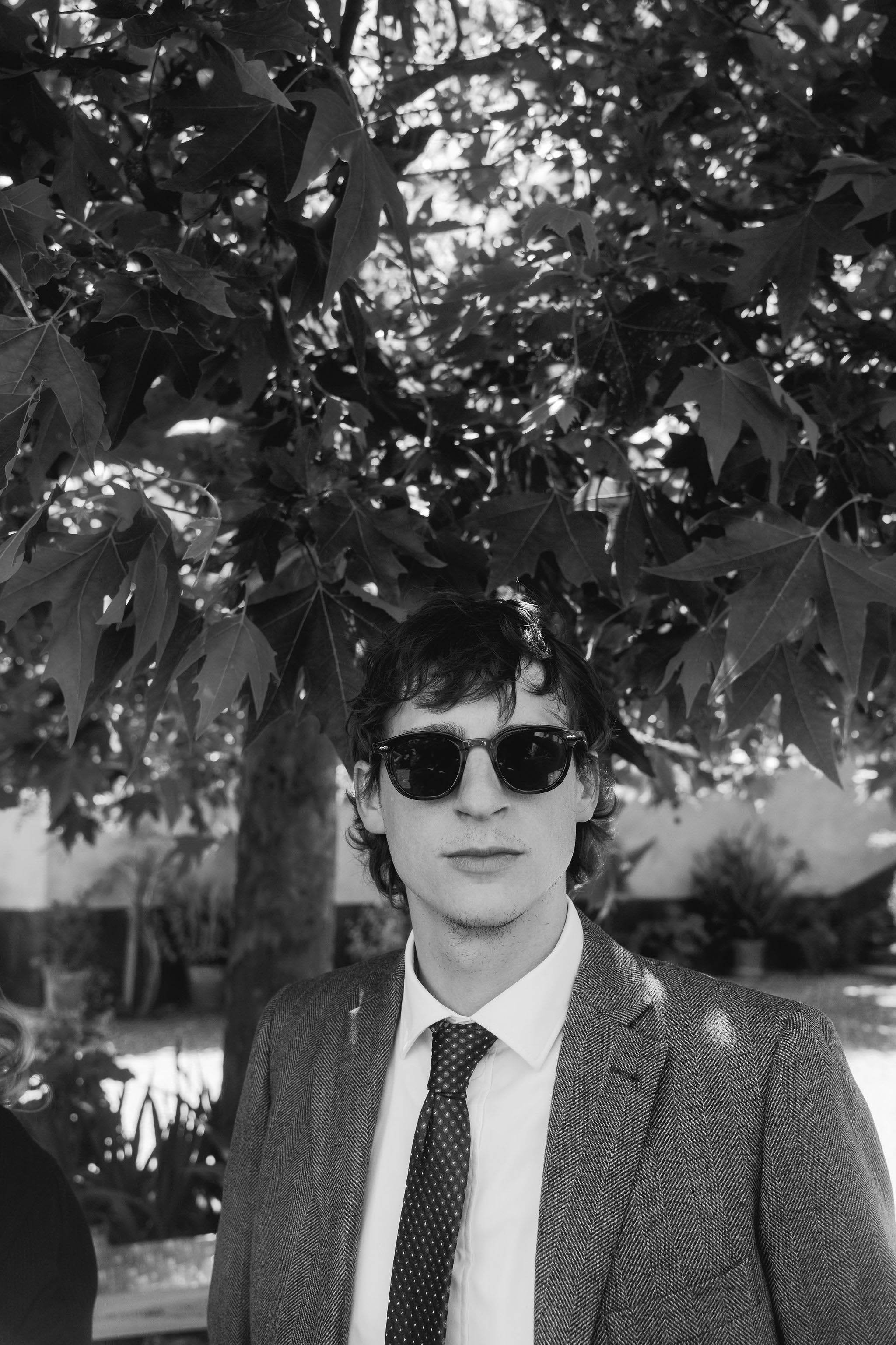 A young man with dark curly hair, wearing sunglasses, a suit jacket, white shirt, and a polka dot tie, standing outdoors under a tree with large leaves.